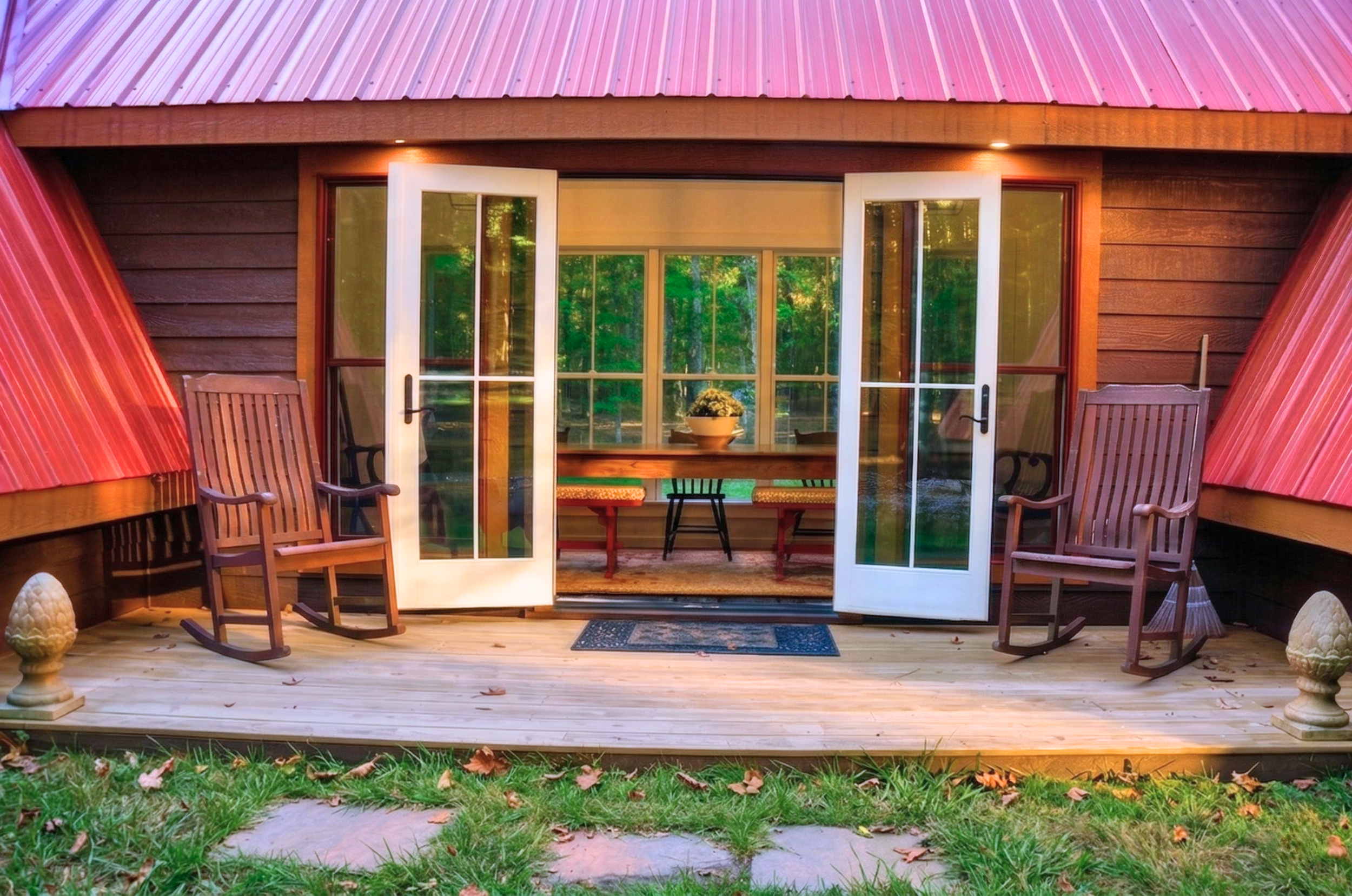 Wooden porch with two rocking chairs, a small black table, and an open glass door leading inside a house with a view of green trees.