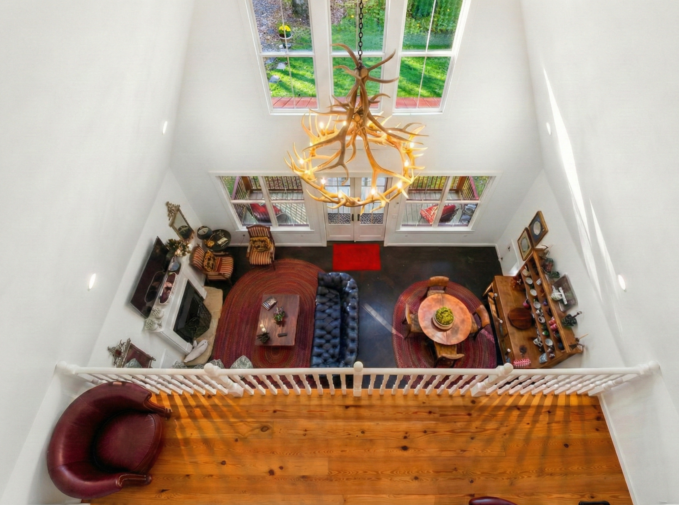 Living room with high white walls and large windows overlooking a green yard. A chandelier made of antlers hangs from the ceiling. The lower level has dark hardwood flooring, a red area rug, a leather armchair, a blue tufted sofa, a wooden coffee tab