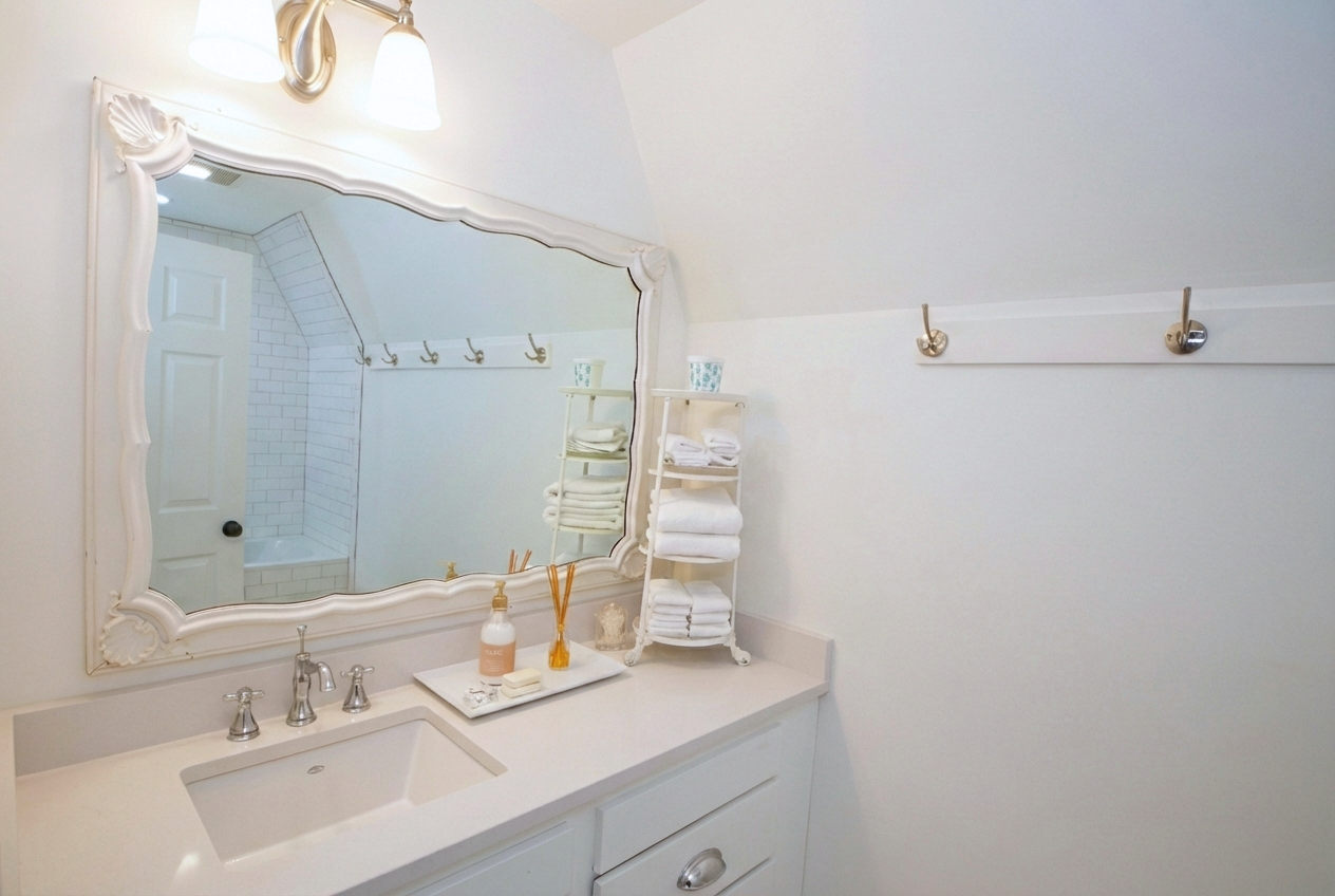 Bathroom with a white sink, large decorative mirror, wall-mounted light fixture, and a small white shelf with folded towels and decorative items. The reflection shows a white door and a corner with white tile and a bathtub.