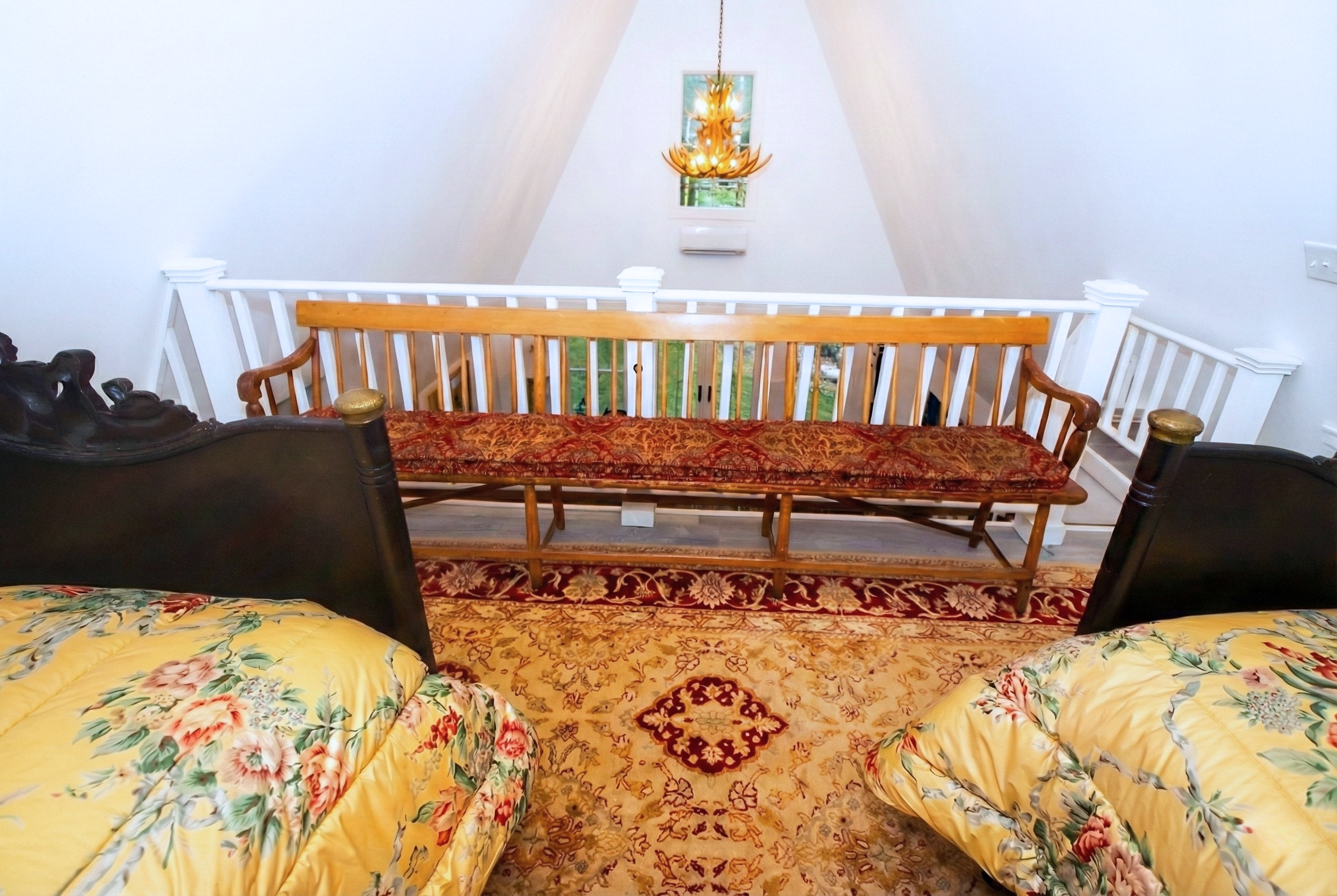 Bedroom with two beds with floral bedding, a red and gold ornate rug, a wooden bench, white and wood railings, a chandelier, and a window with a view outside.