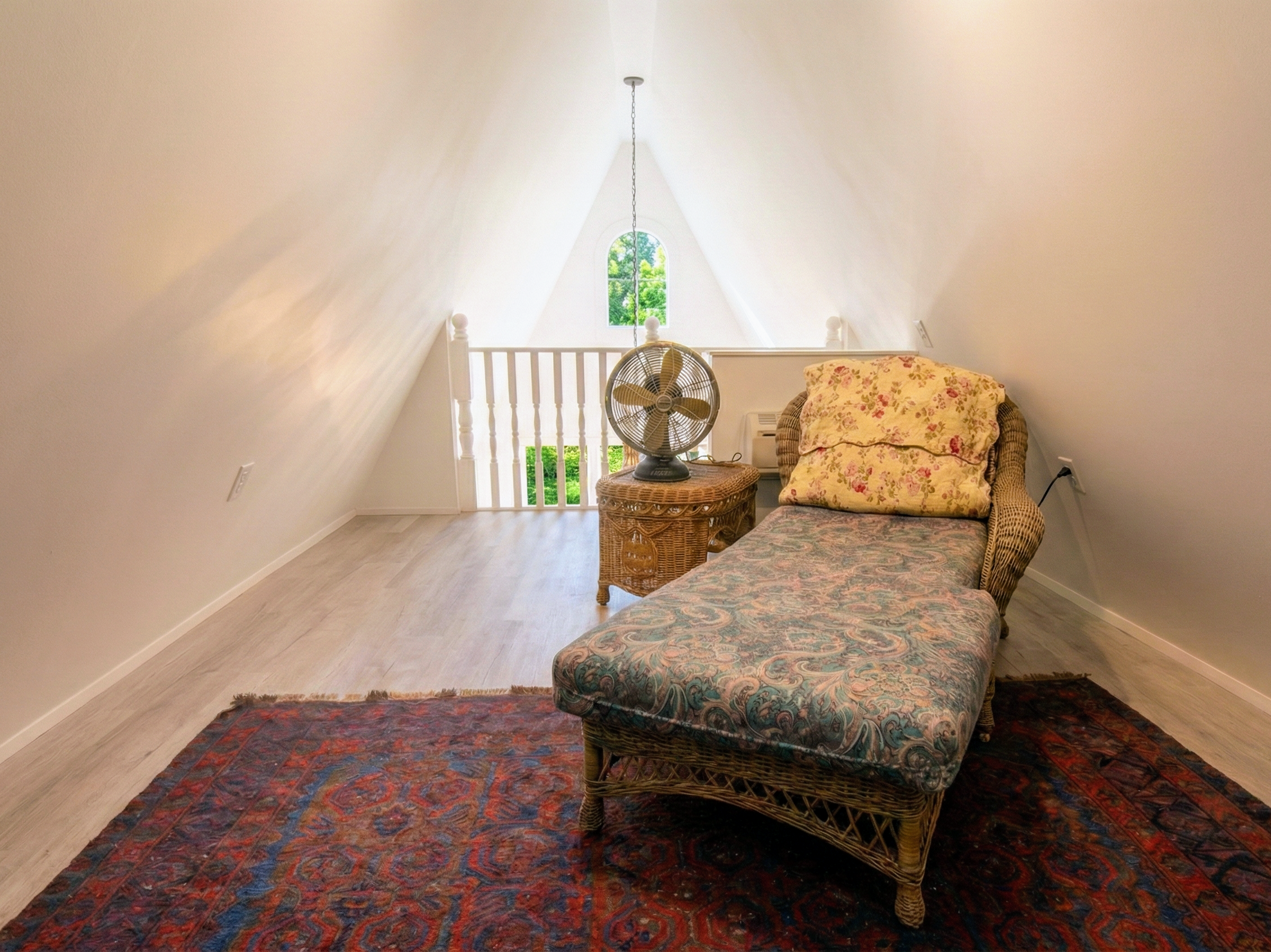 A cozy attic room with sloped white walls, a small window, a wicker chaise lounge with floral cushions, a wicker side table, a vintage fan, and a colorful rug on light wood flooring.