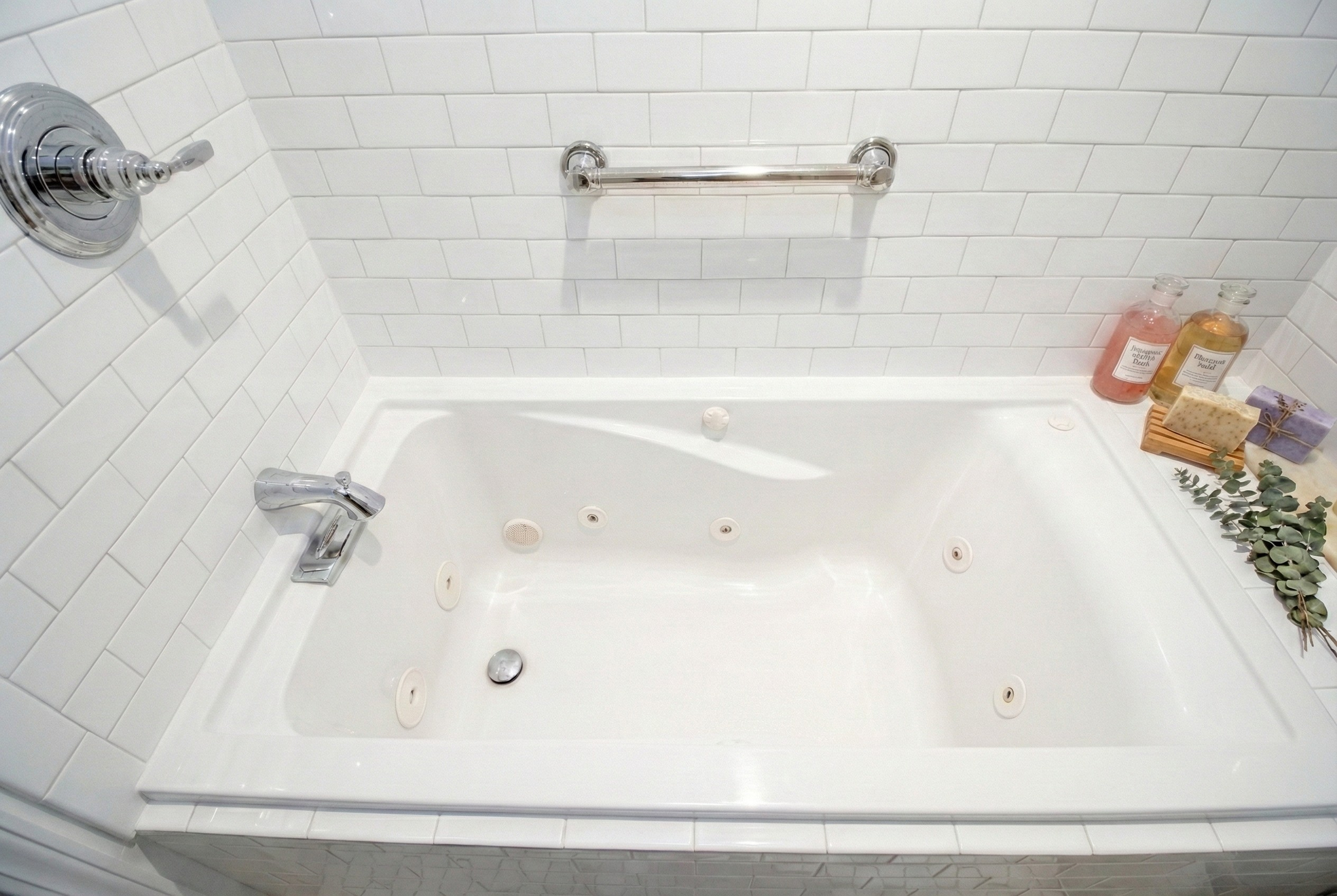 A white bathtub with multiple jets, a silver faucet, and a wall-mounted grab bar in a tiled bathroom corner, with bottles of bath products, soap, and a sprig of eucalyptus on the edge.