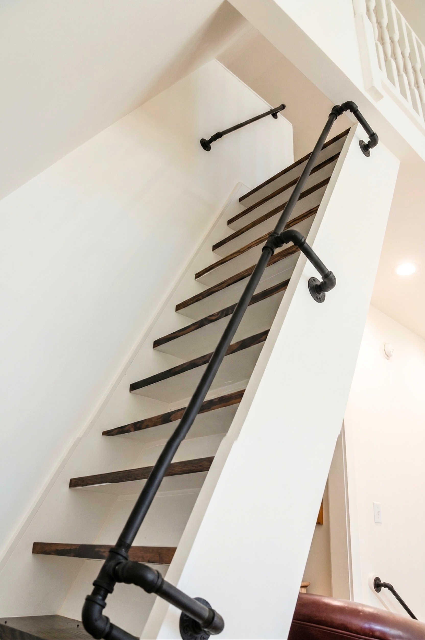 View of a staircase with wooden steps and black pipe railing, leading upward to a first-floor landing with a white railing, in a modern home interior.