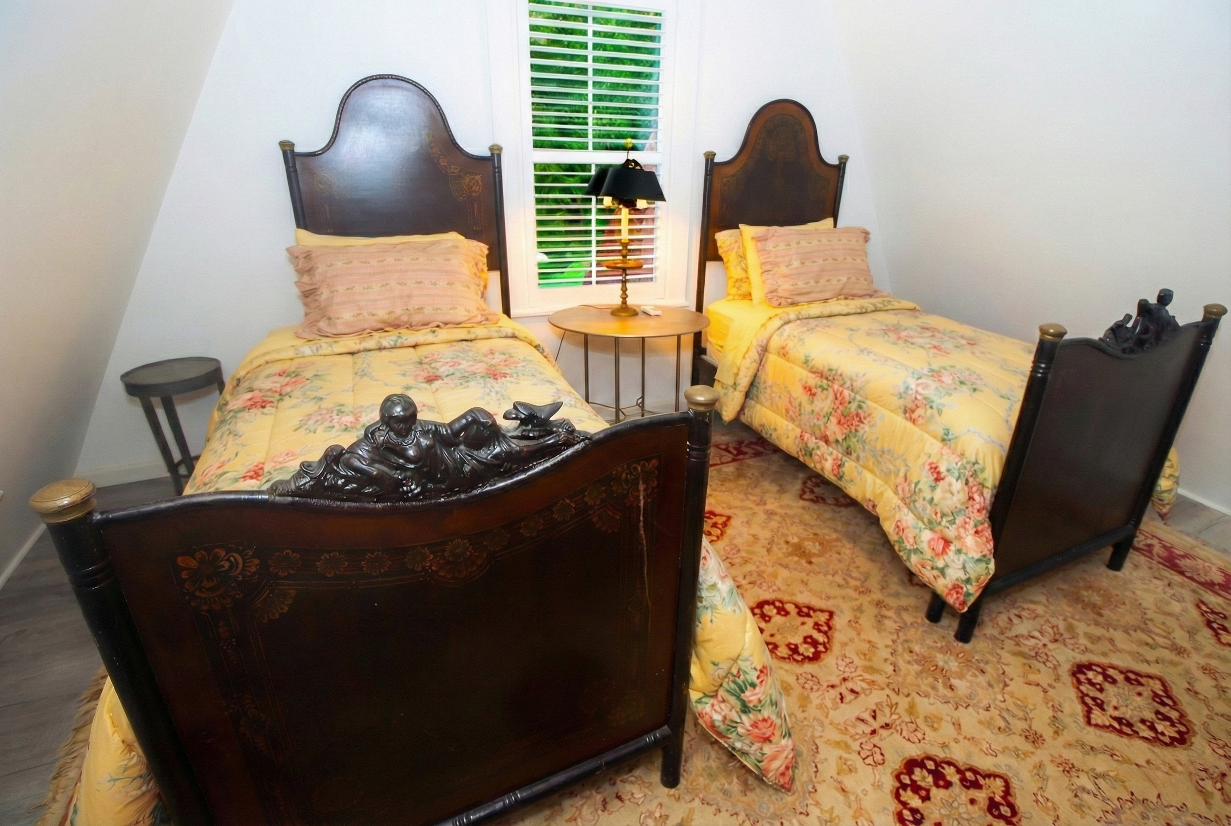 A bedroom with two single beds with dark wooden headboards and footboards, floral bedding, and pink pillows. There is a small round table with a lamp between the beds, a window with blinds behind it, and a patterned area rug on the floor.