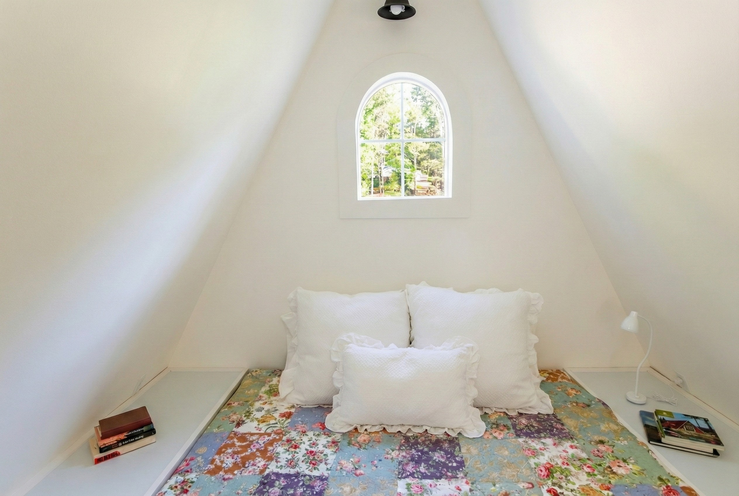 A cozy bedroom with white walls and a quilted bedspread with a floral pattern. Three large white pillows are arranged against the wall. There is a small window with an arched top showing trees outside. Two white side tables hold books and a white des