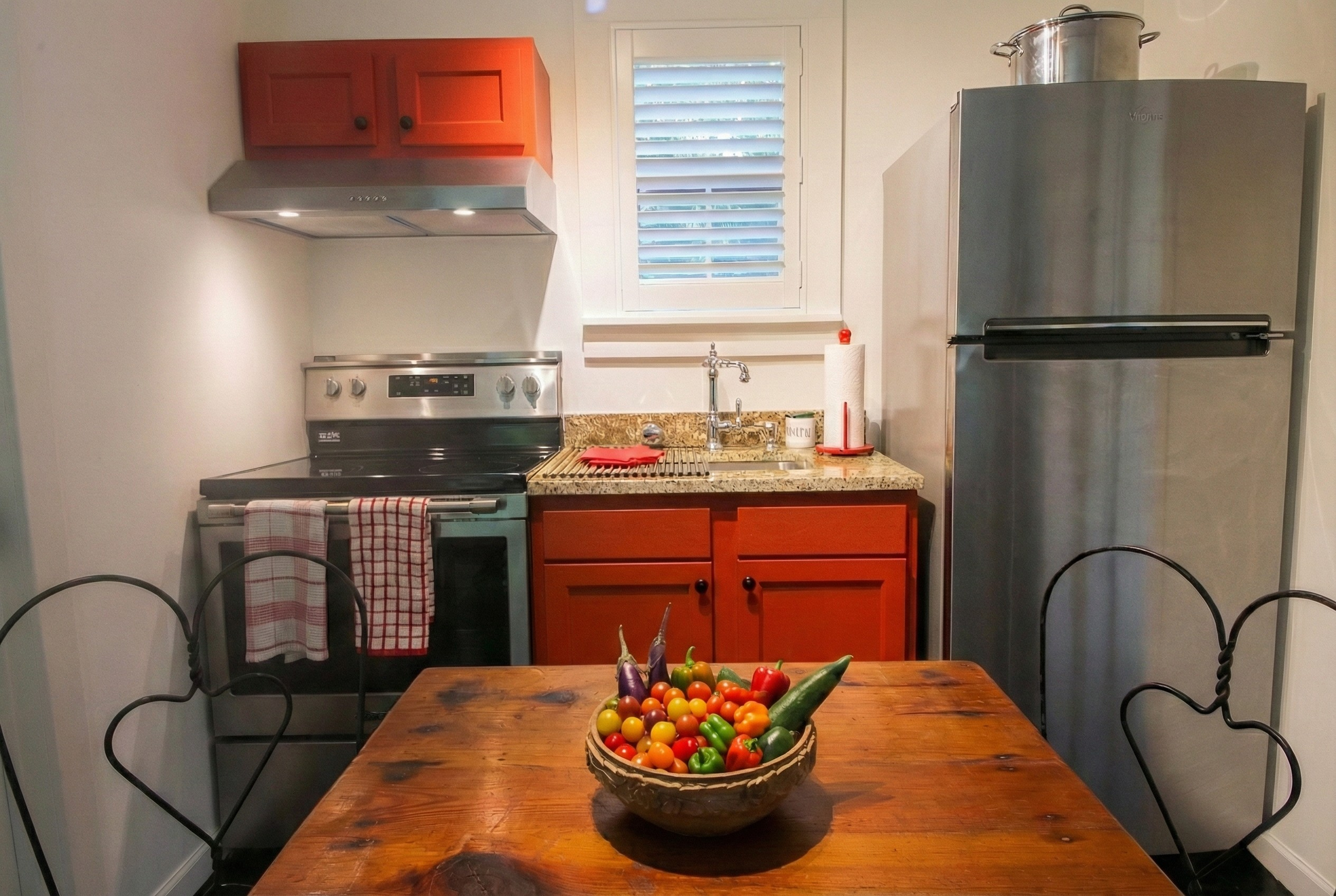 A kitchen with a wooden dining table, a bowl of colorful vegetables, a stove with towels, a granite countertop with a sink, a window with shutters, a red cabinet, and a metallic refrigerator.