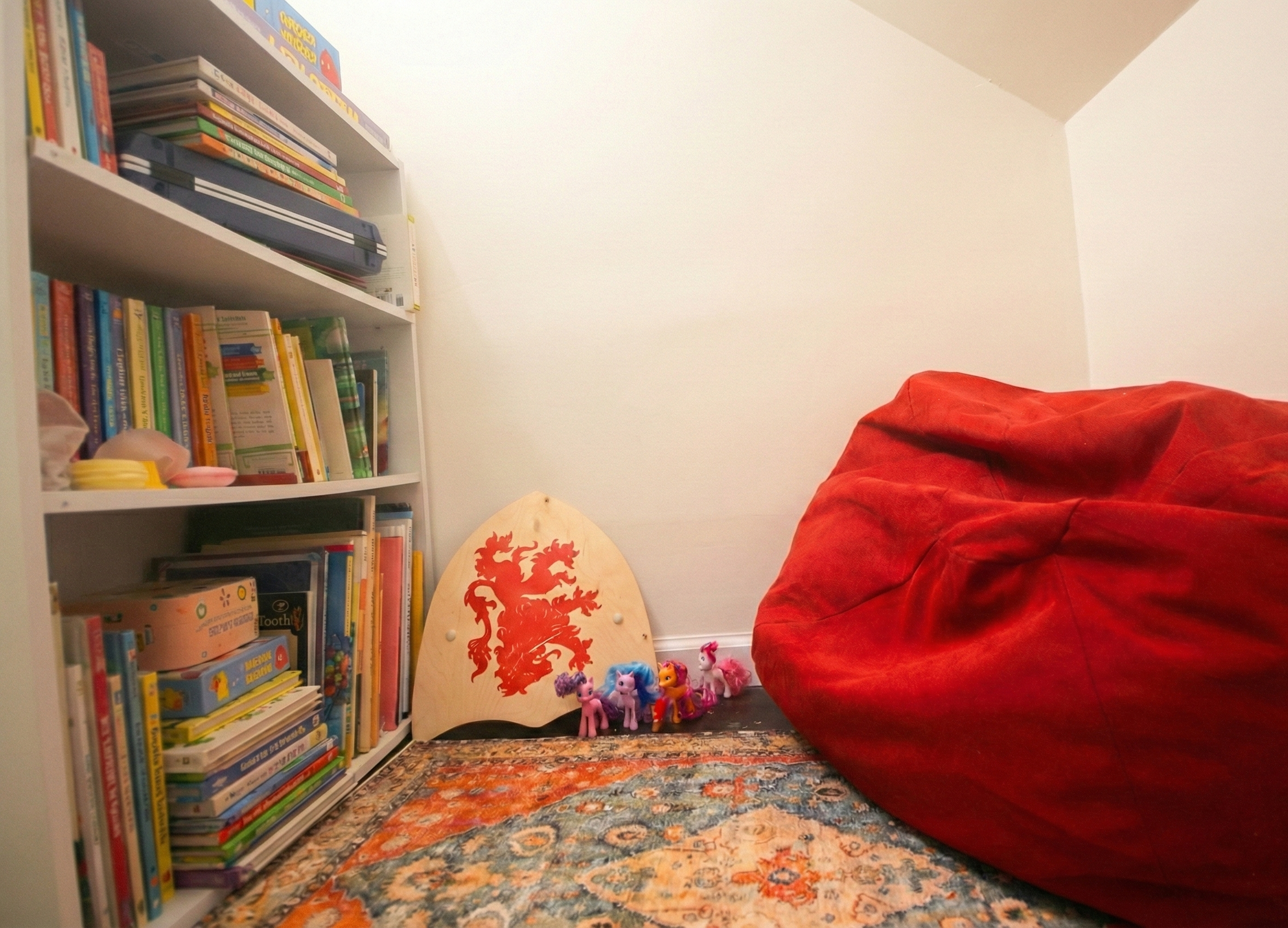 A corner of a room with a white bookshelf filled with colorful books on the left side. Next to it, on the floor, there is a wooden shield with a red lion crest on it, and several small toy ponies with colorful manes lined up in front. On the right si