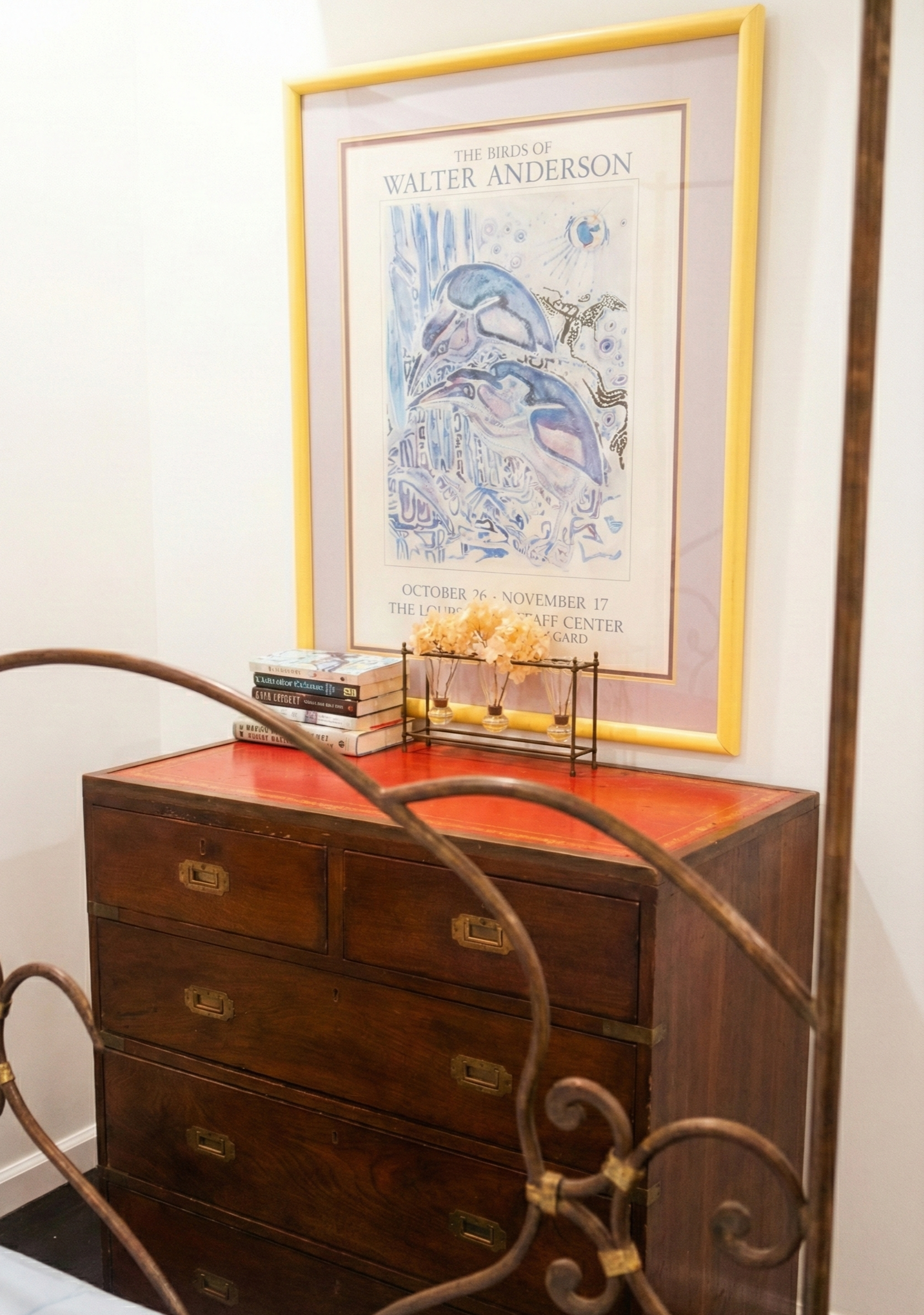 A vintage wooden dresser under a large Walter Anderson art poster with yellow border, featuring watercolor-style bird illustrations, next to a small decorative table with books and dried flowers, viewed partially through a wrought iron railing and mi