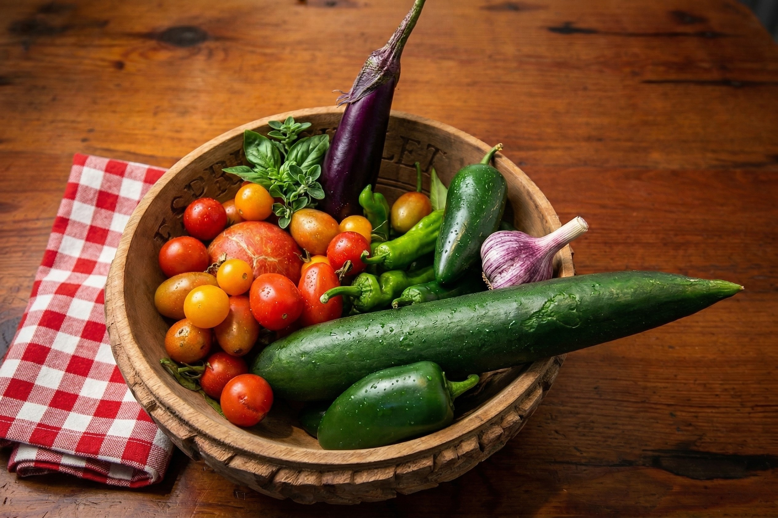 Fresh vegetables in a wooden bowl on a wooden table, including cherry tomatoes, a cucumber, eggplant, garlic, and chili peppers, with a red and white checkered napkin nearby.