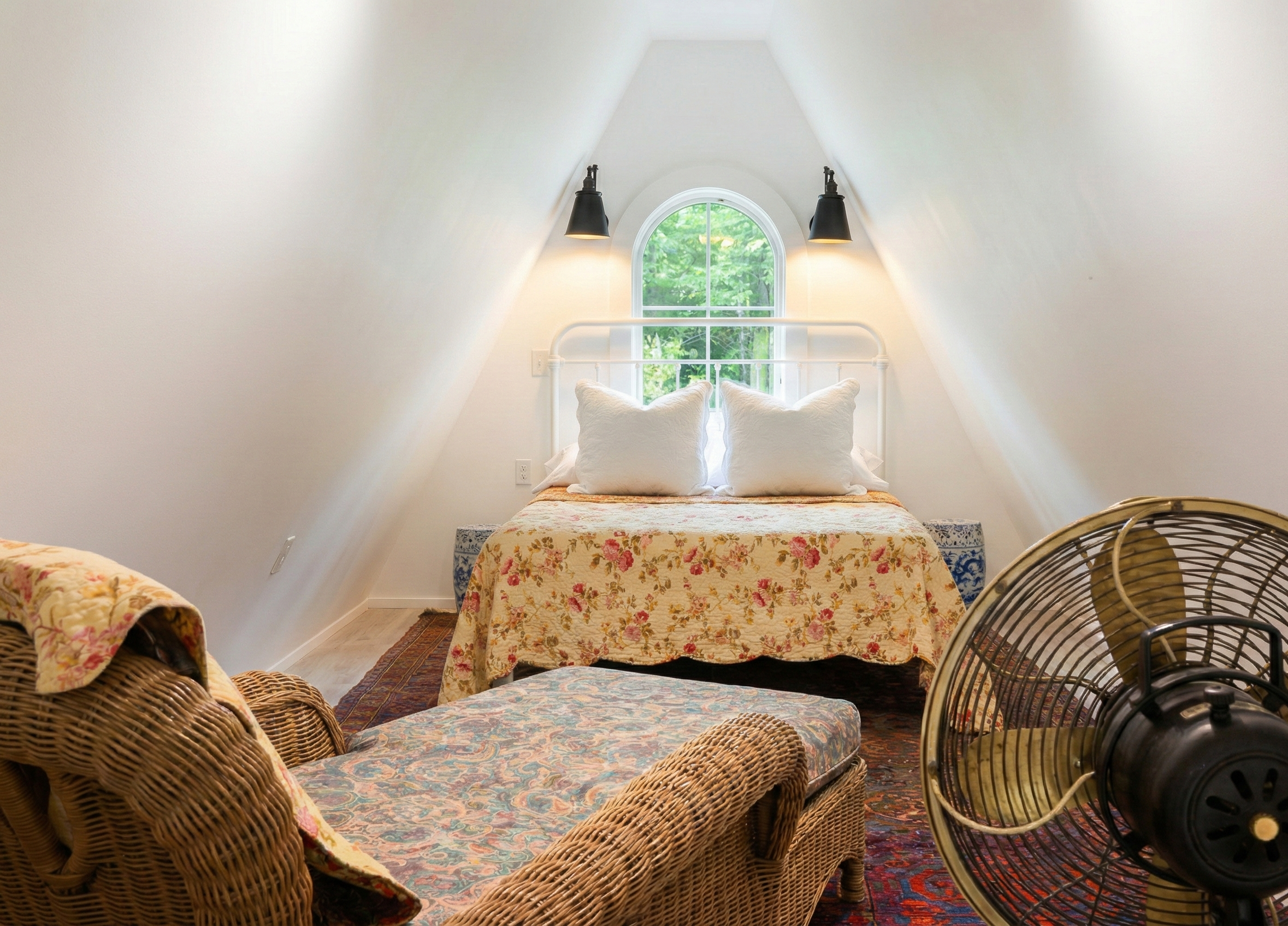 Cozy attic bedroom with white walls and slanted ceiling, featuring a white bed with floral quilt and pillows, a wicker chair with a floral cushion, a vintage rug, a window with greenery outside, black wall sconces, and a black fan.
