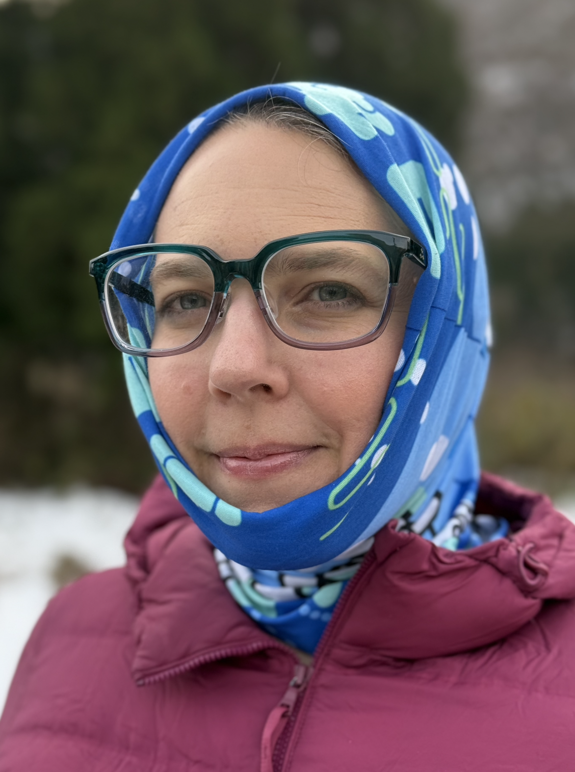 A product image of a ski scarf pulled over the head of a female model standing in a snow. The Japanese souvenir is blue, teal, white and black illustrative image of a family of kimono dressed kokeshi and daruma doll snowboarder and skiers.