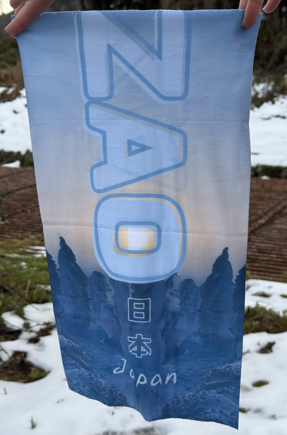 Image of a rectangular shape tube scarf featuring the Zao Snow monsters at dusk in a pale blue with the sun setting above in a pale yellow. Text reads Japan and Zao.