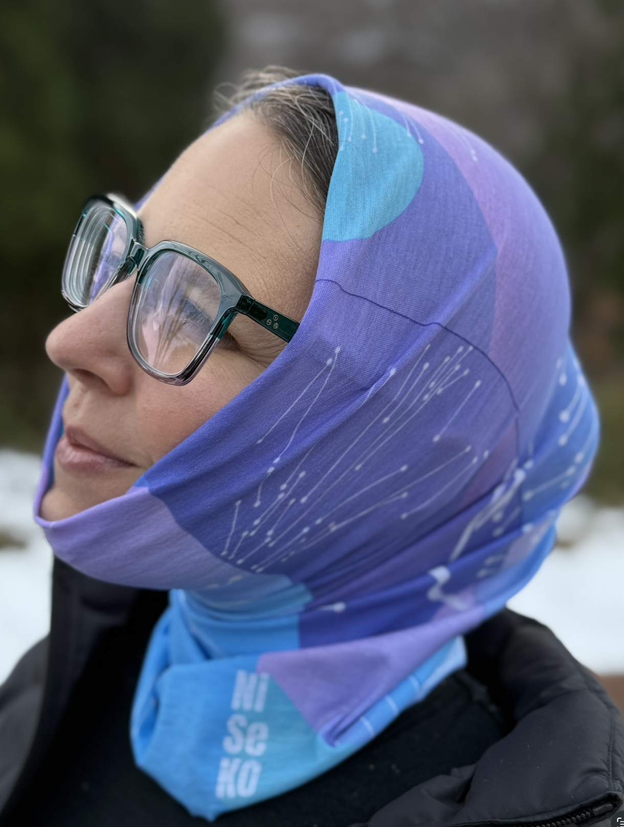 Female wears blue and purple tube scarf overhead. Scarf has lines that look like constellations and the word Niseko at the bottom.