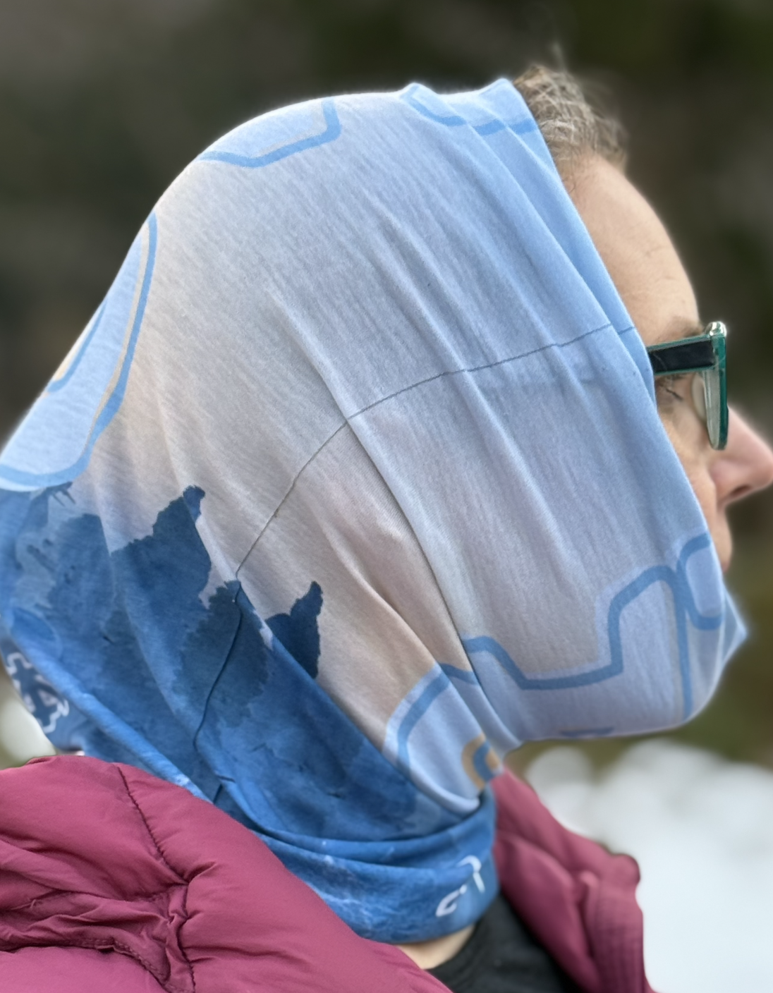 Woman wearing a tube scarf pulled over her head. The scarf is blue and light yellow featuring a picture of snow covered trees known as snow monsters.