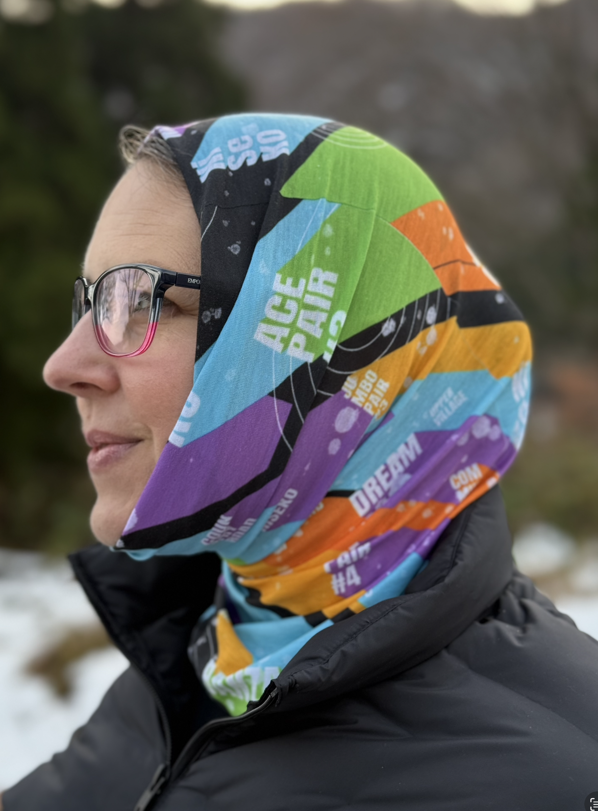 Ski scarf pulled over the head of a female. Scarf has the pattern of colorful blocks with the names of ski lifts in 3D block typography with subtle line art detail.