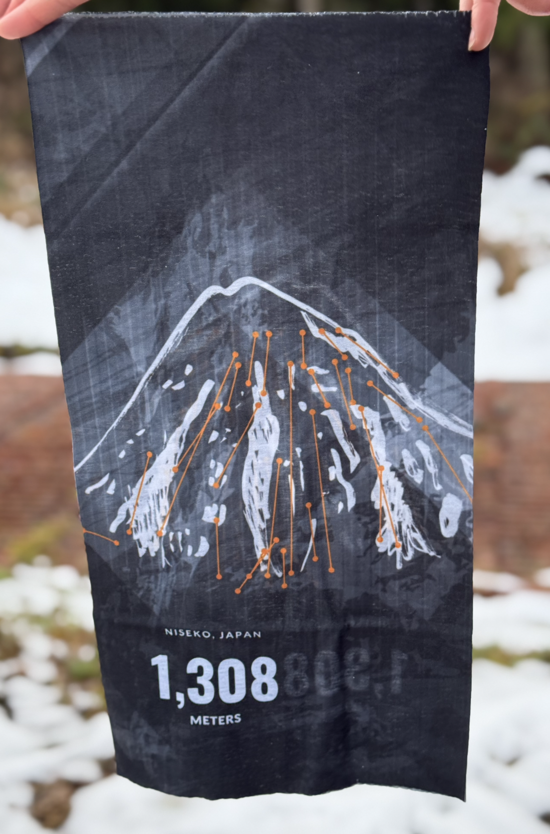 Black, orange, and white tube scarf held up in a winter environment. Design is a calligraphy mountain with orange lines that represent the ski lifts.