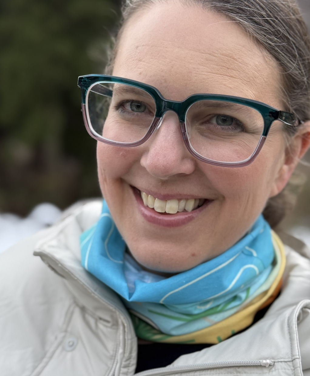 A rectangular tube scraf with a blue to orange gradation, japanese pine trees, and daruma dressed as snowboarder or skiiers with Japanese Kanji for Japan and snow. Scarf is modeled by a female and bunched around the neck.