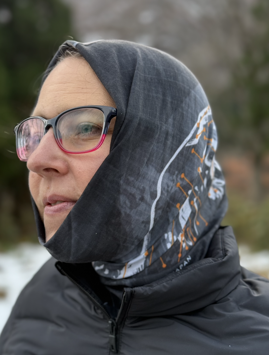 Black, orange, and white tube scarf pulled over the head of a female. Design is a calligraphy mountain with orange lines that represent the ski lifts.