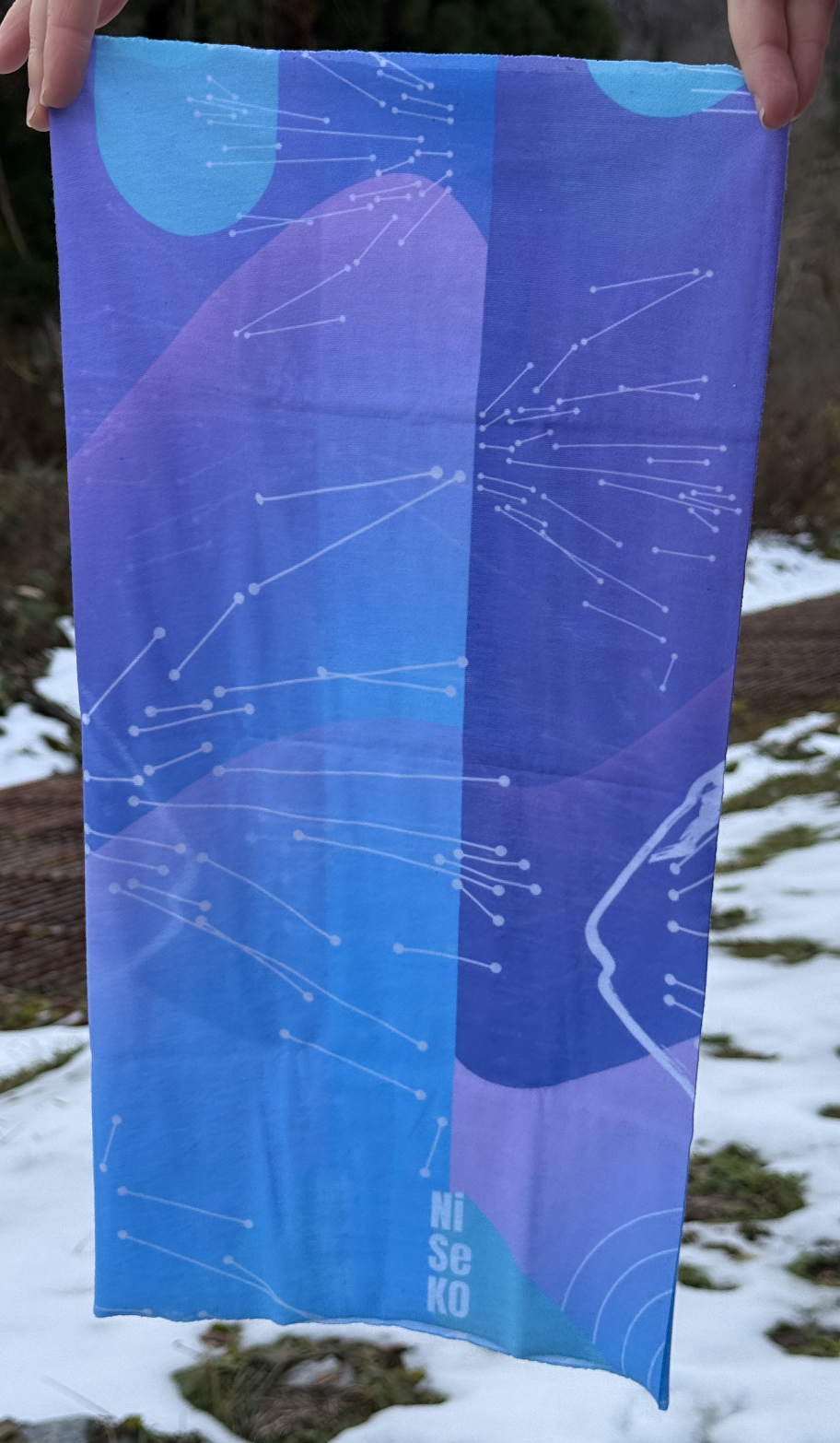 Blue and purple rectangular scarf held up in a winter environment. Scarf looks like it is a constellation map, but is a map of the resort lifts.