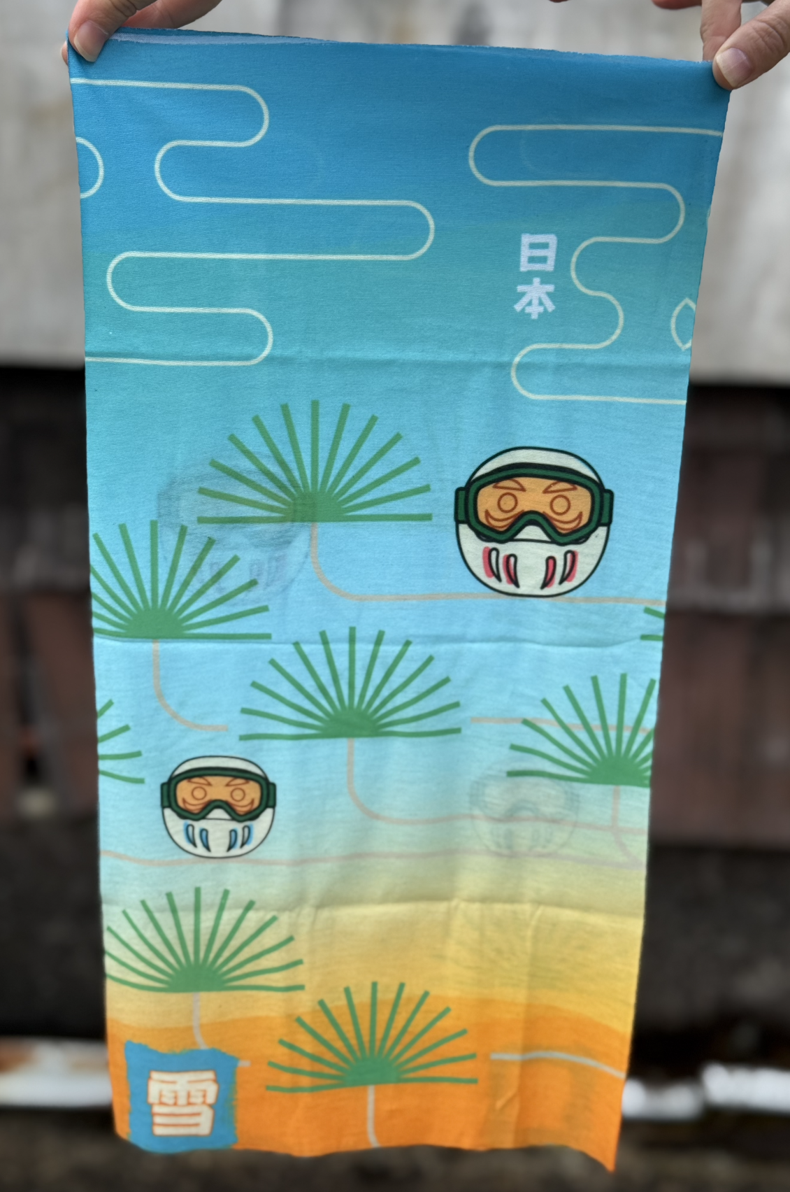 A rectangular tube scraf with a blue to orange gradation, japanese pine trees, and daruma dressed as snowboarder or skiiers with Japanese Kanji for Japan and snow.