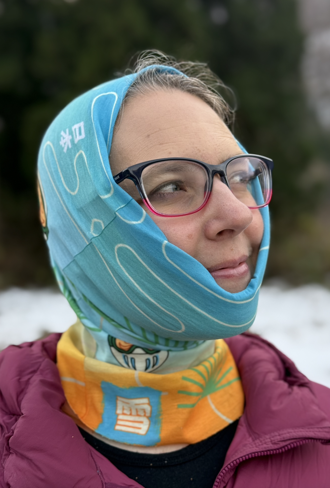 A rectangular tube scraf with a blue to orange gradation, japanese pine trees, and daruma dressed as snowboarder or skiiers with Japanese Kanji for Japan and snow. Scarf is modeled by a female with the scarf over her head.