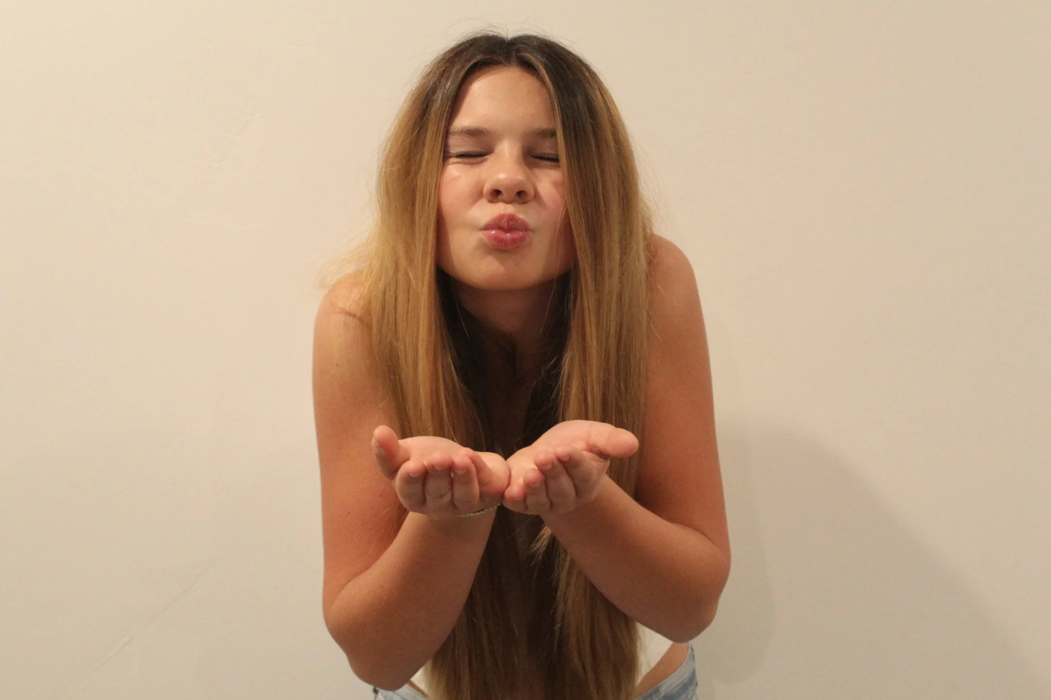 A young woman with long, light brown hair blowing kisses with her eyes closed, standing against a plain white wall.