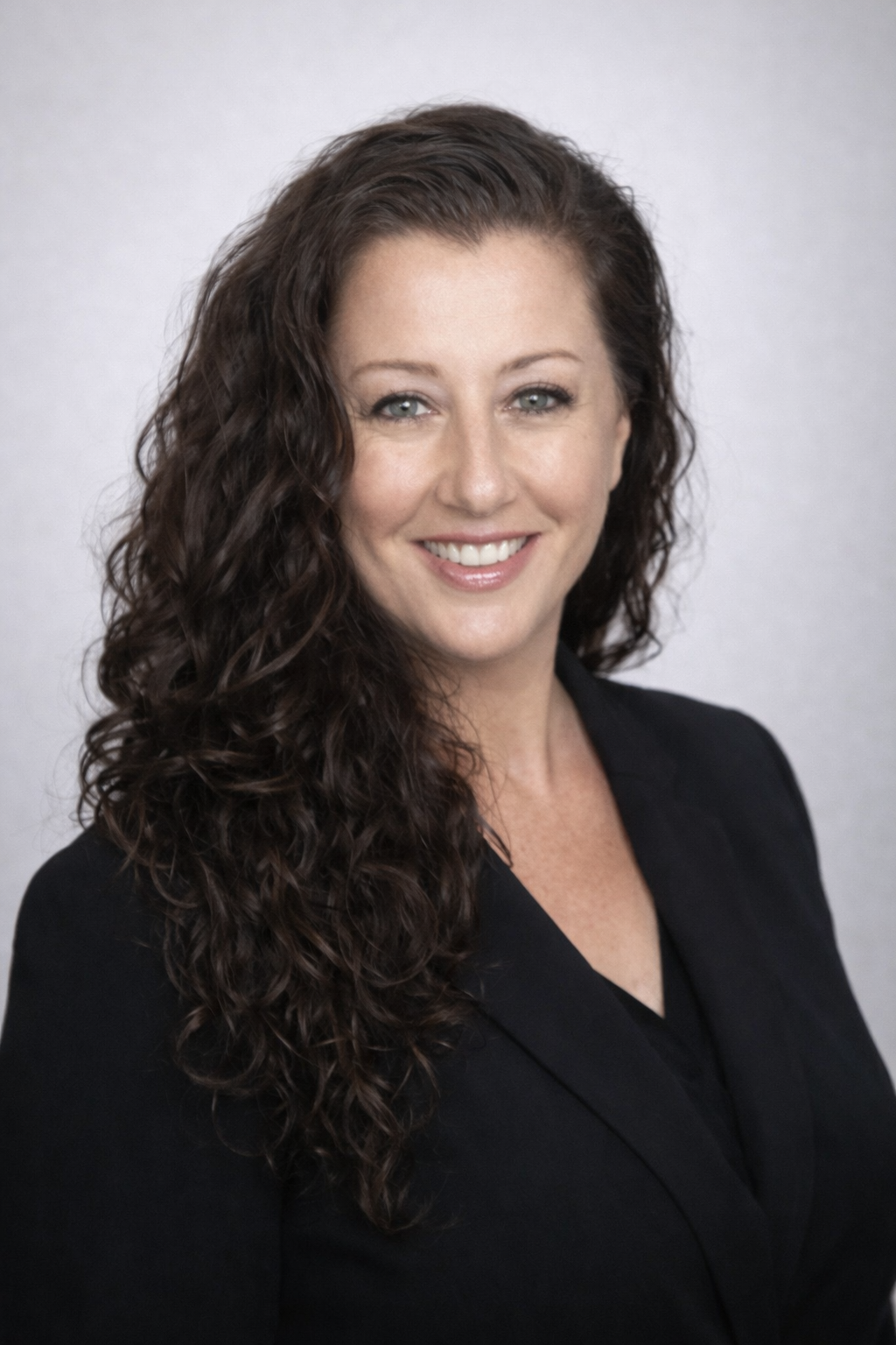 Professional headshot of a woman with long, curly dark hair, wearing a black blazer and smiling.