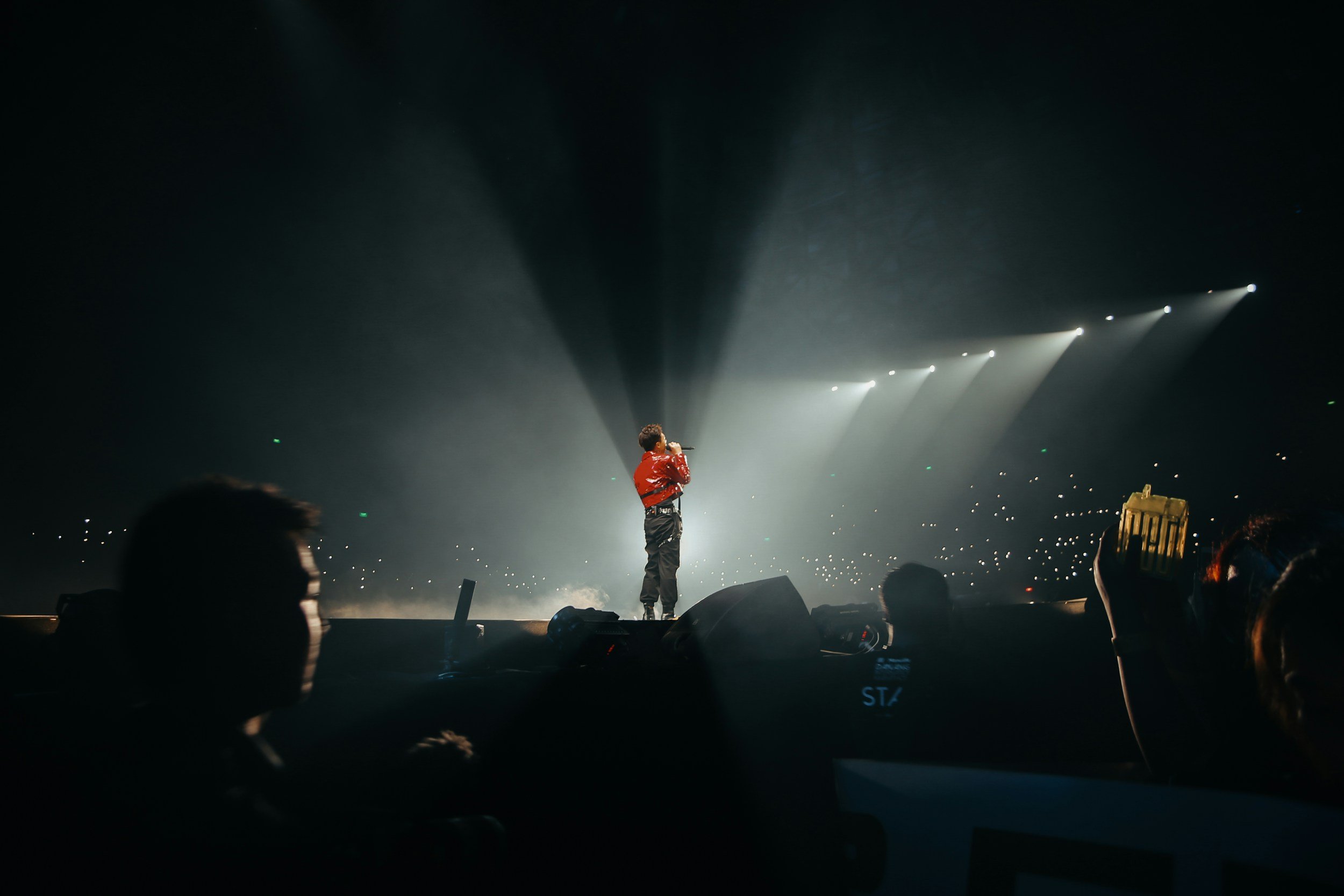 A singer in a red jacket performing on stage during a concert, with bright lights and an audience in the background.