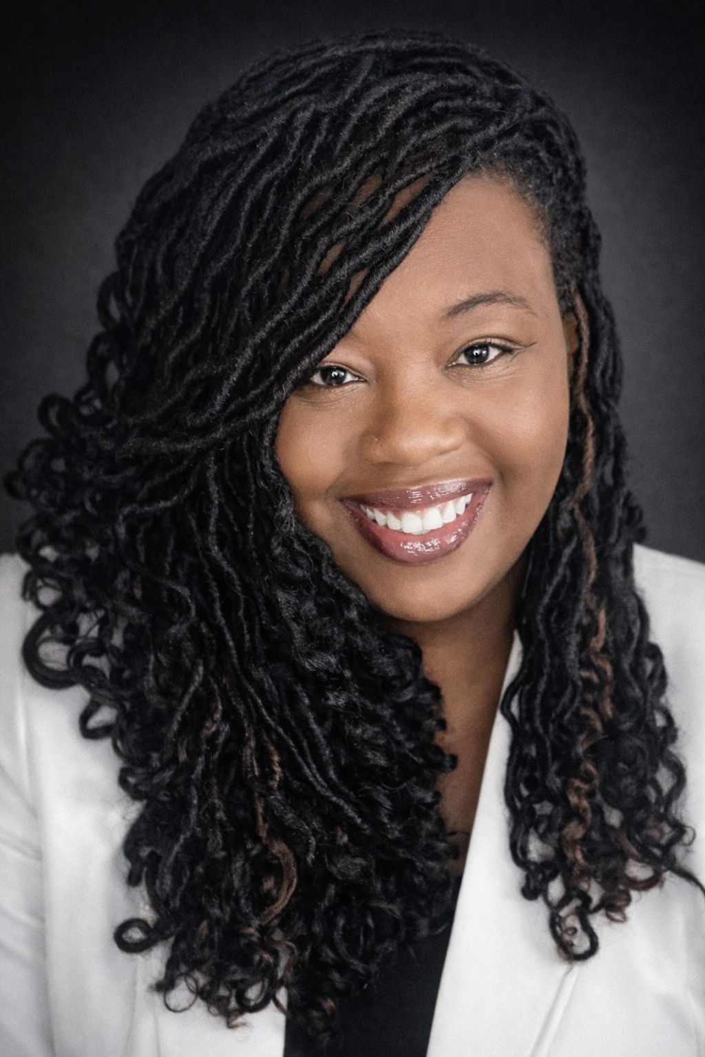 Close-up portrait of a smiling woman with dark, curly, shoulder-length hair, wearing a white blazer and dark lipstick against a black background.