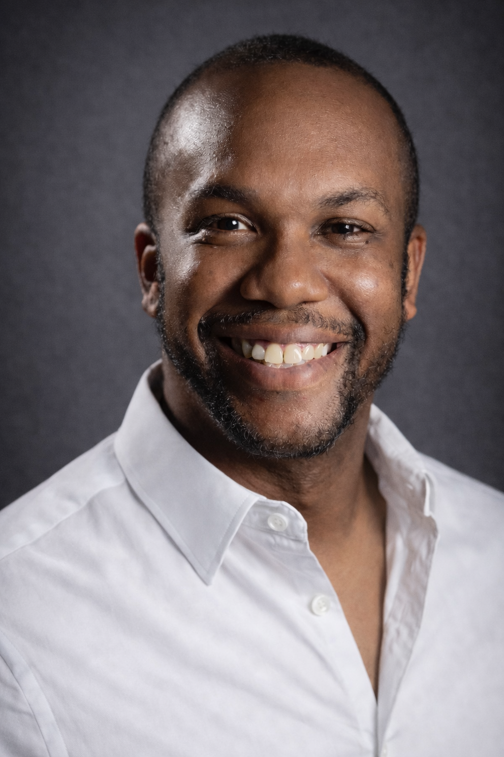 Close-up of a smiling African American man with short hair and a beard, wearing a white button-up shirt, against a dark background.