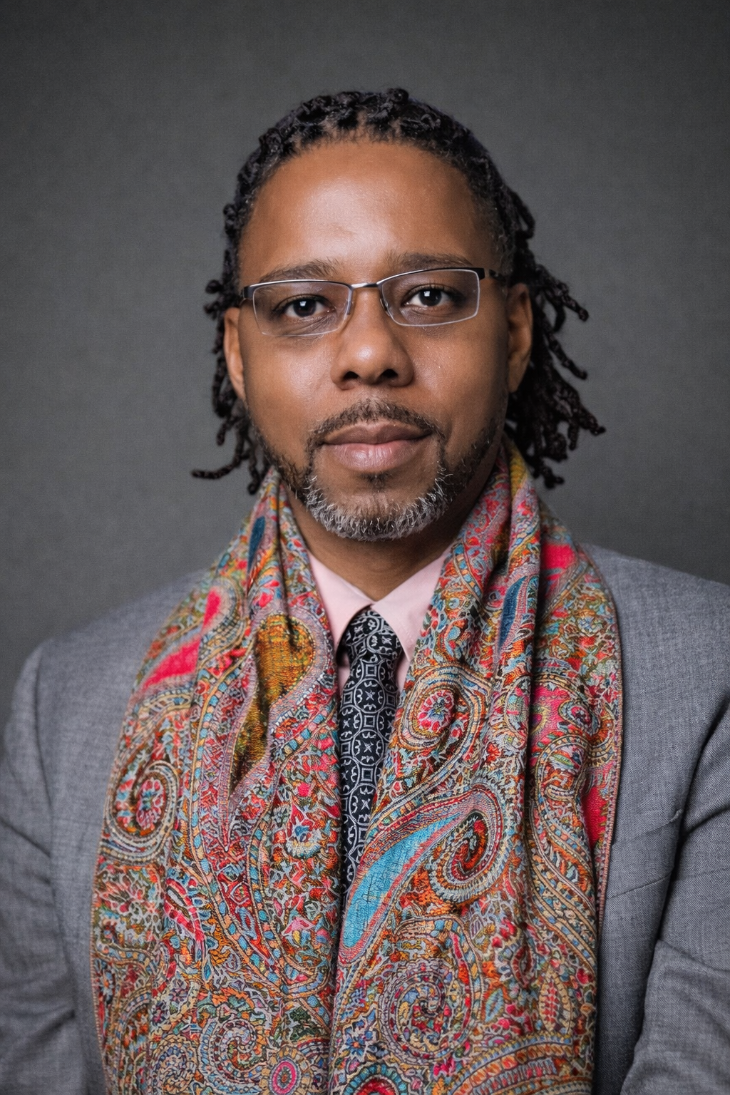 A man with glasses, a gray suit, a pink shirt, a black patterned tie, and a colorful paisley scarf, posing against a dark gray background.