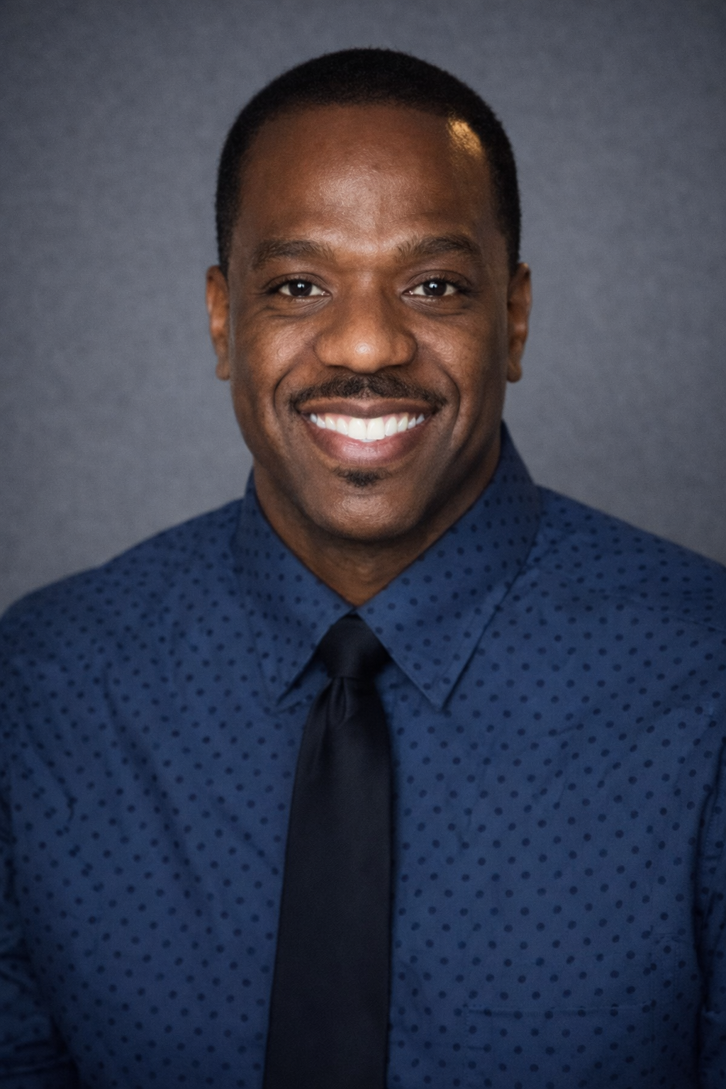 A smiling man with short black hair, wearing a dark blue shirt with small black polka dots and a black tie, standing against a gray background.