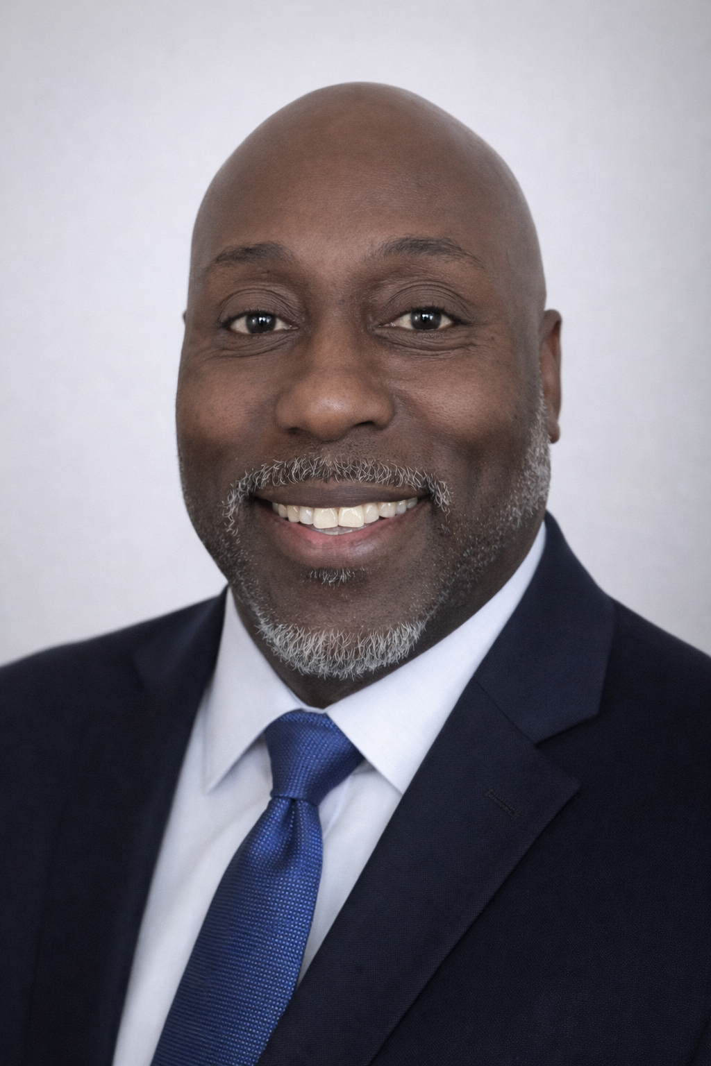A professional headshot of a smiling African American man wearing a navy suit, white shirt, and blue tie, against a plain light background.