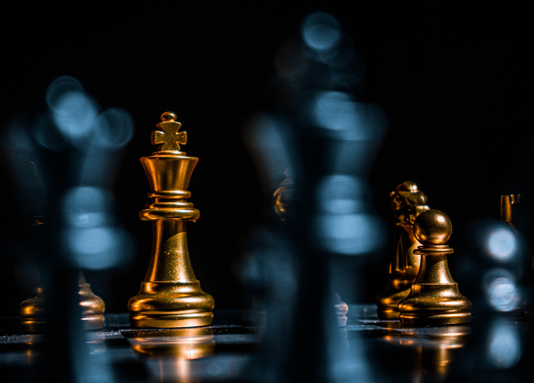 Close-up of gold chess pieces, including a queen and a pawn, on a chessboard with a dark background and bokeh lighting.