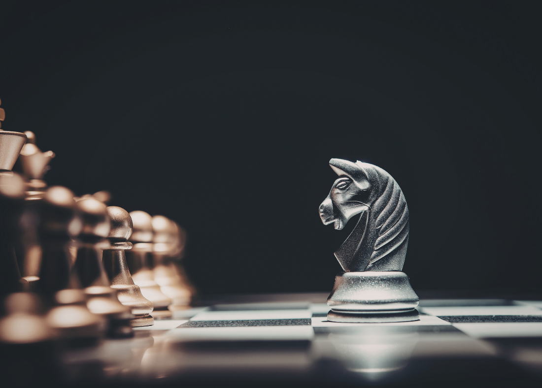 Close-up of a metallic chess knight piece facing the camera on a chessboard, with other pieces blurred in the background.
