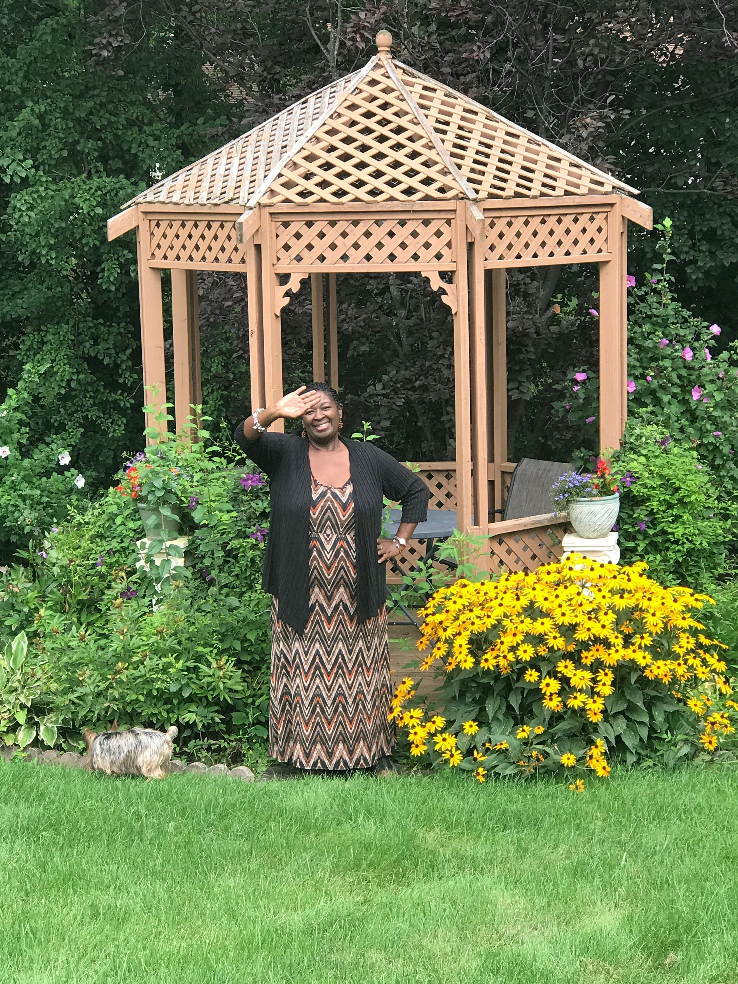 A woman standing in a garden with a wooden gazebo, surrounded by colorful flowers and a small dog at her feet, smiling and waving.