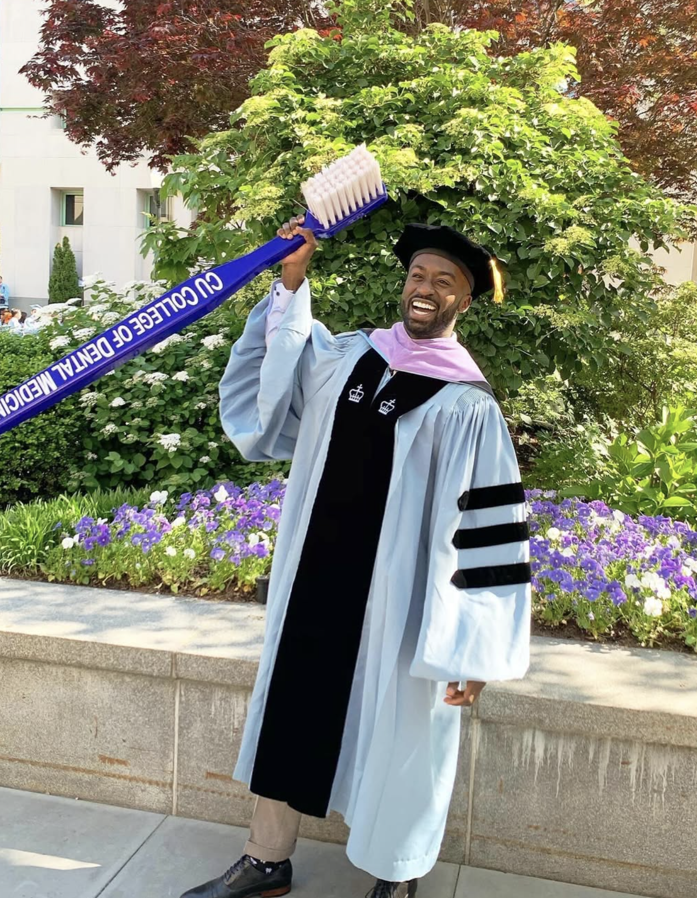 A man in graduation cap and gown holding a giant blue diploma-shaped prop that says 'CU COLLEGE OF DENTAL MEDICINE' while smiling outdoors in front of green bushes and flowers.