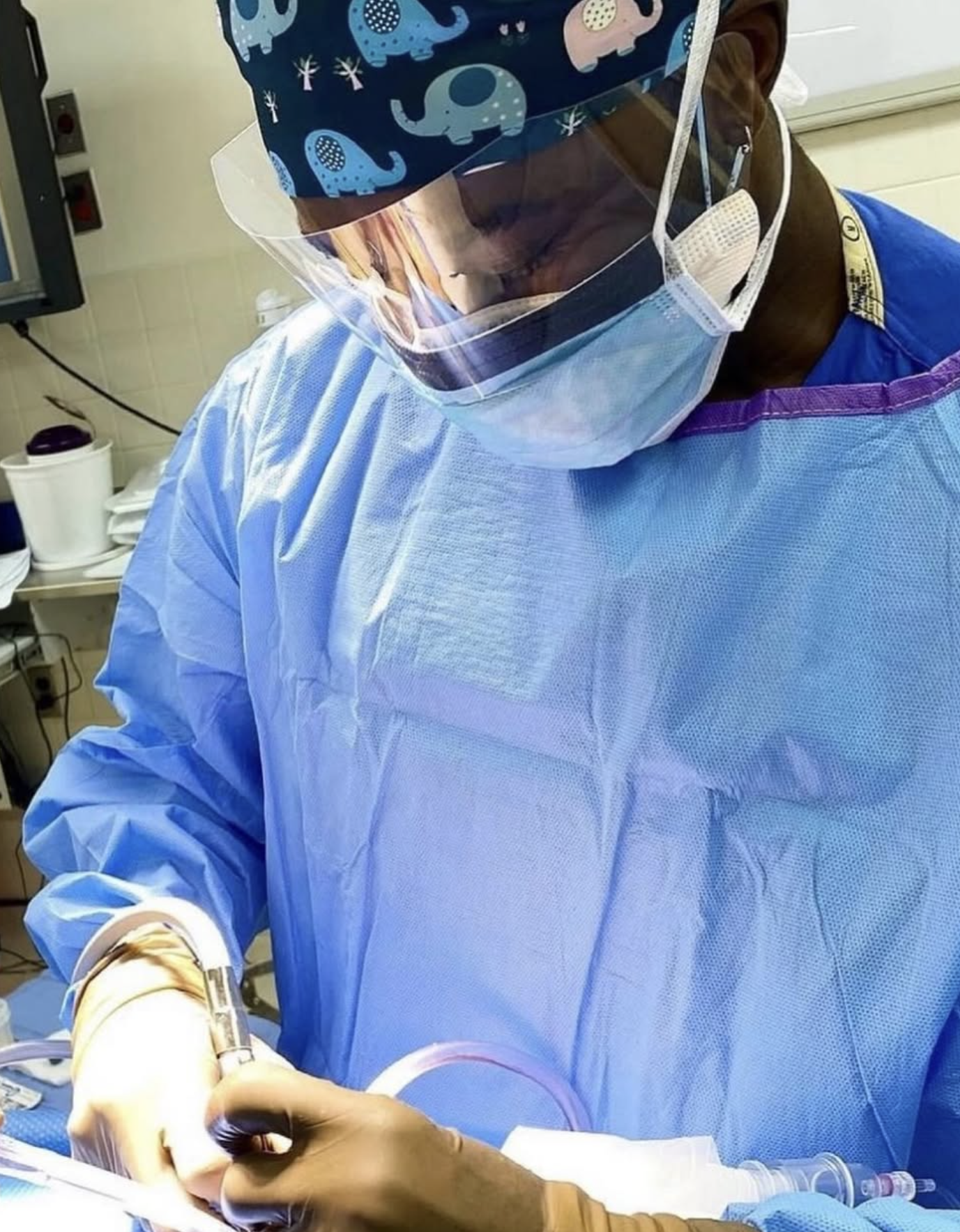 A healthcare professional dressed in blue protective gown, wearing a face shield, face mask, and surgical cap with elephants, is performing a medical procedure in a clinical setting.