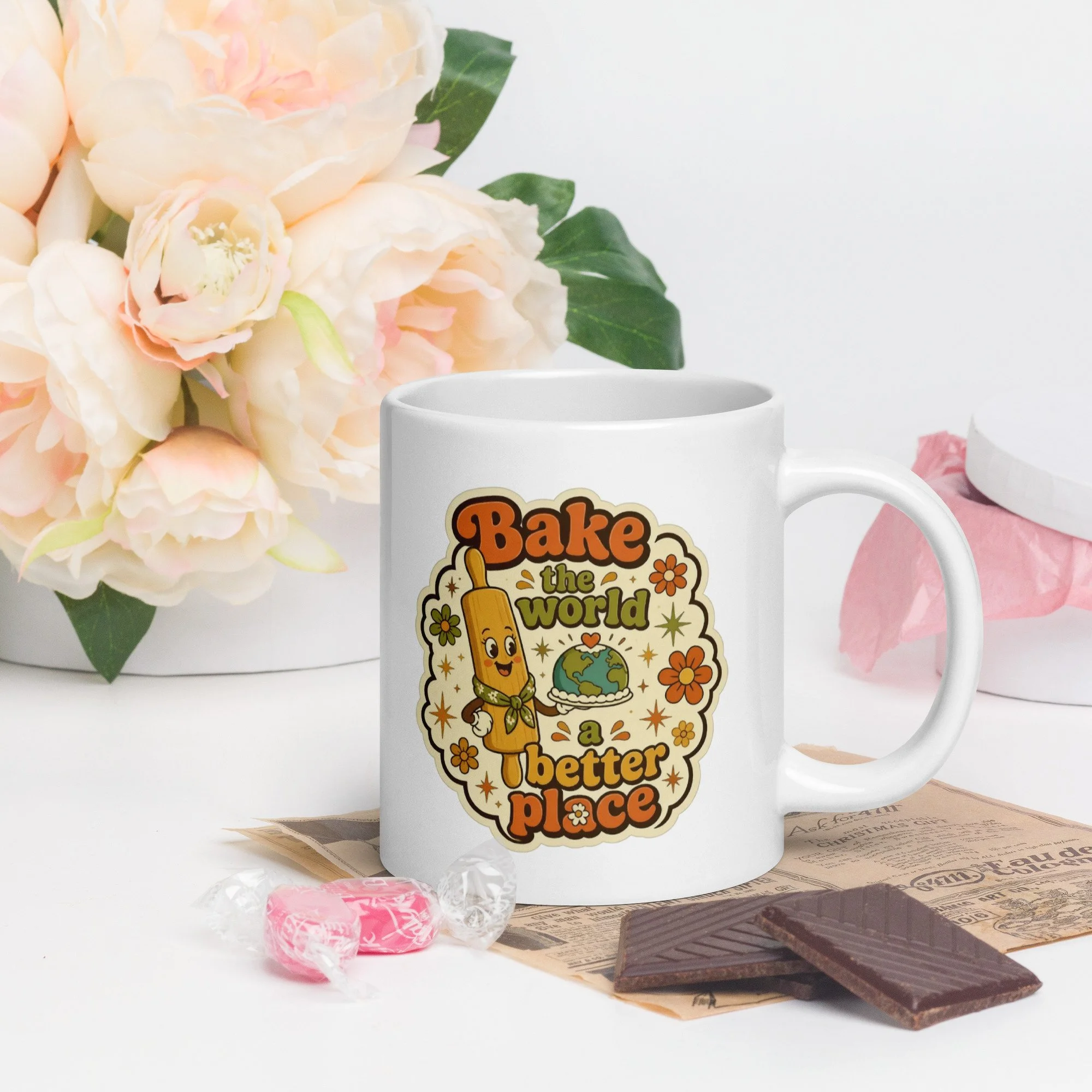 Styled photo of the retro baking mug with “Bake the world a better place” design on a table with flowers and baking props.