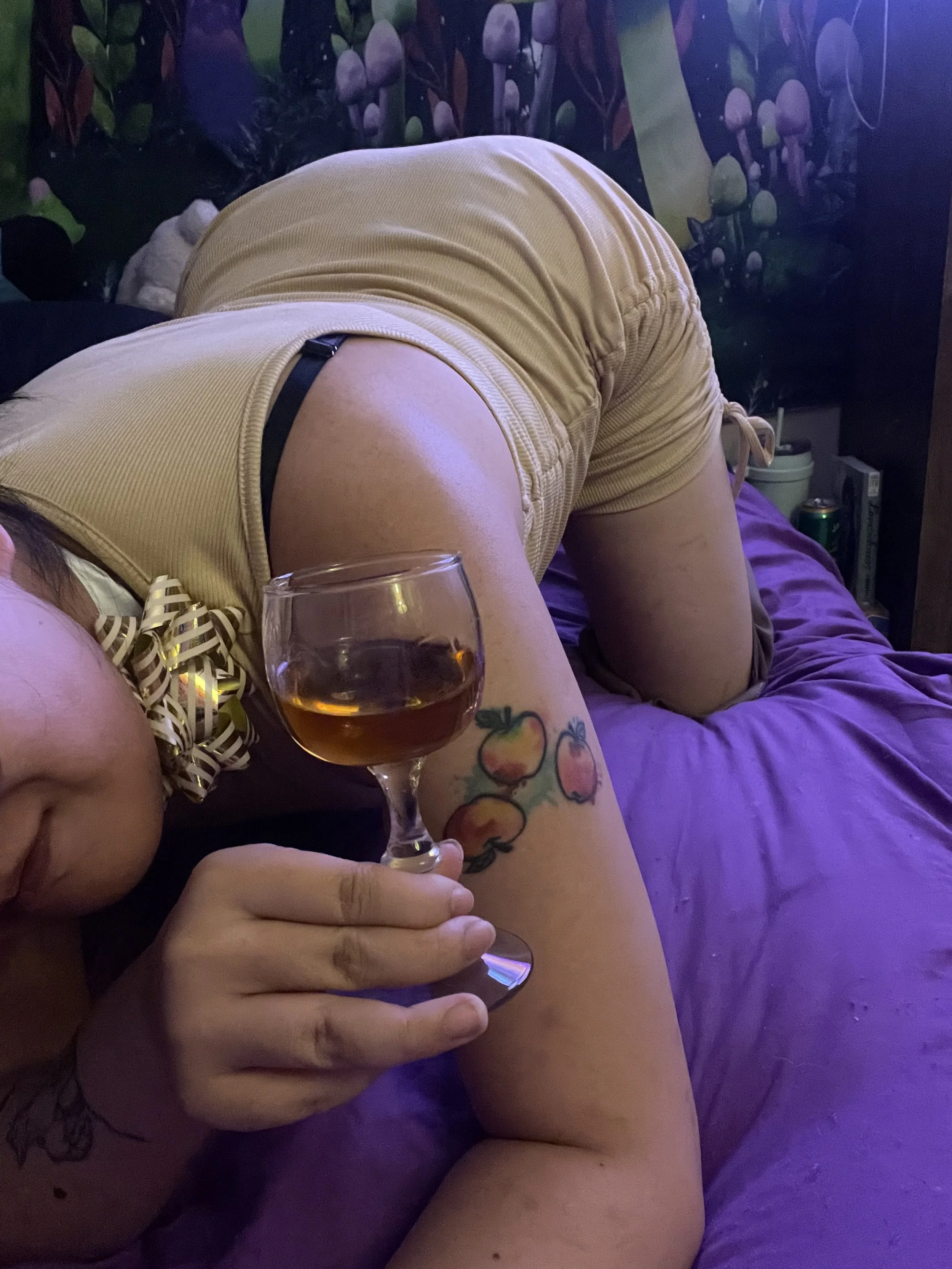 A person lying on their side on a bed, holding a glass of wine, with a colorful tattoo of peaches on their arm. The person is wearing yellow shorts and a matching yellow top with a bow. The background features a mushroom-themed wall decoration.