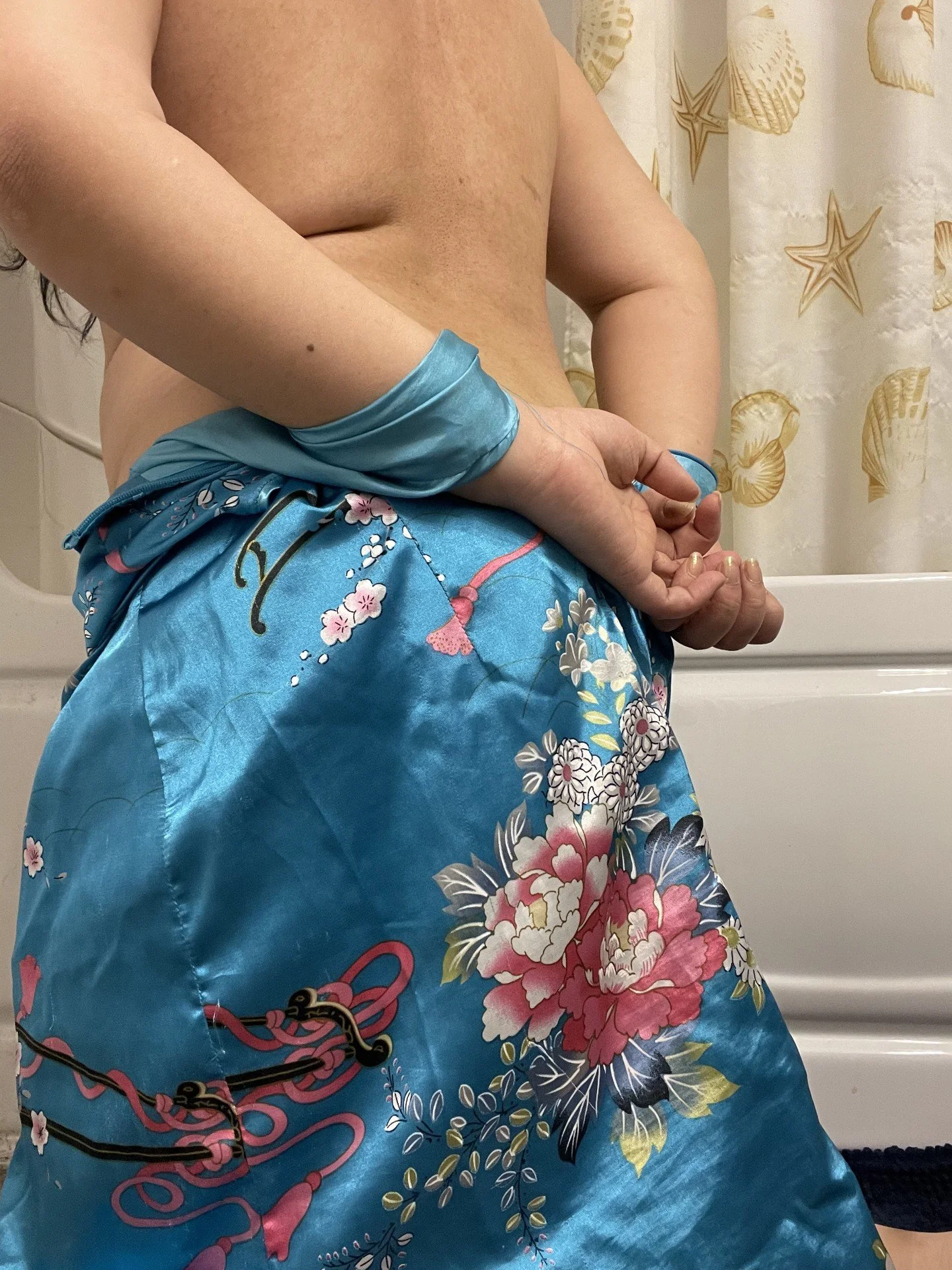 A person standing in a bathroom with floral and seashell curtains, wearing blue silk shorts with a pink and white floral pattern, and a matching blue silk wristband.