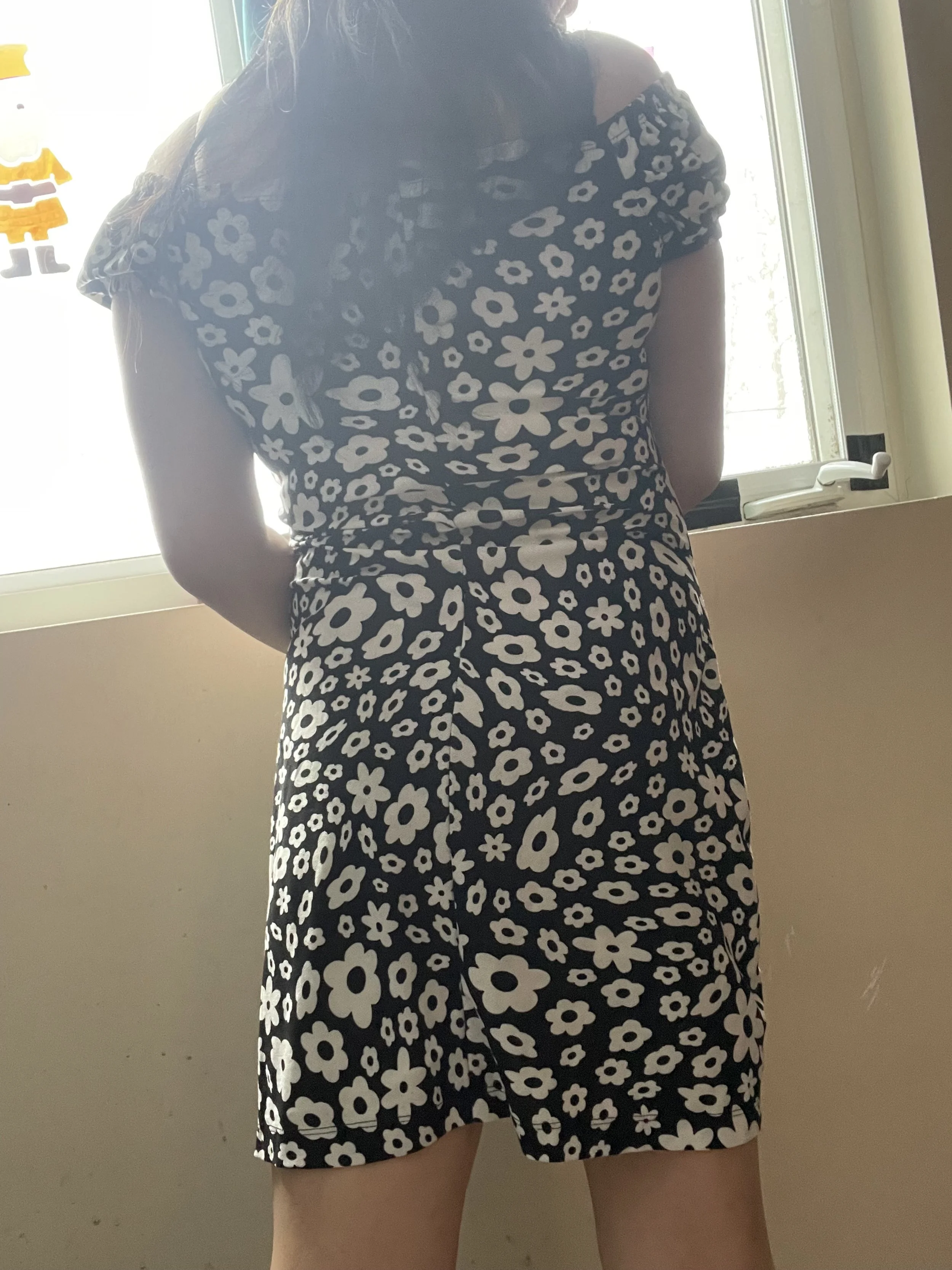 Back view of a woman wearing a black dress with white floral pattern, standing in front of a window.