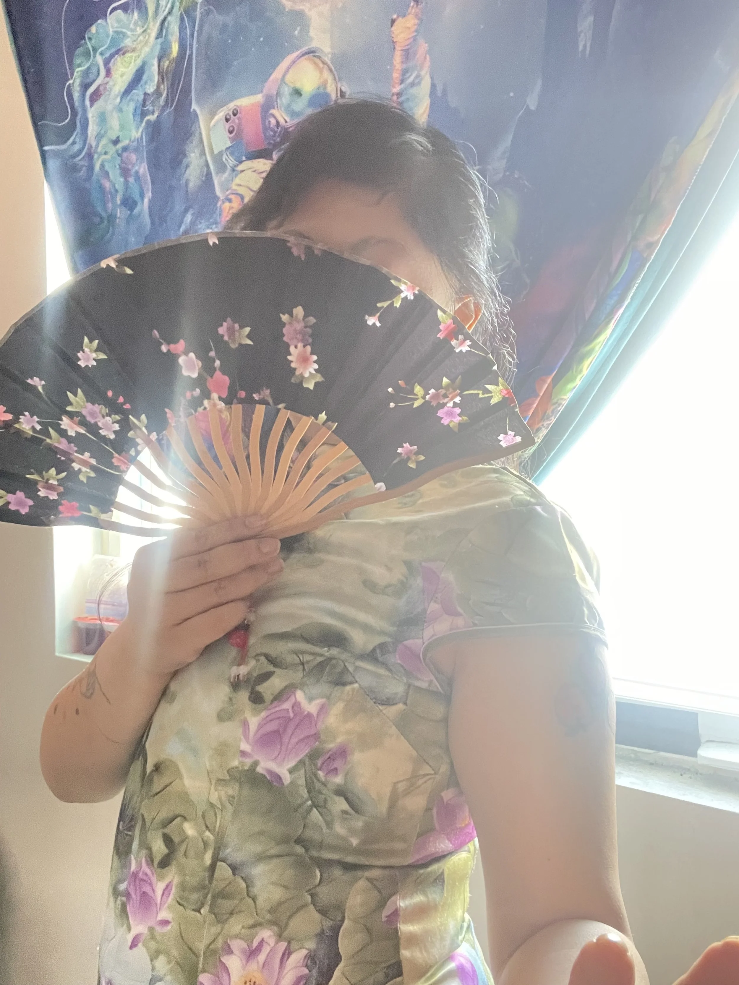 Person holding a decorated black fan with pink and purple flowers, partially covering their face, standing in front of a colorful space-themed poster, with sunlight coming through a nearby window.