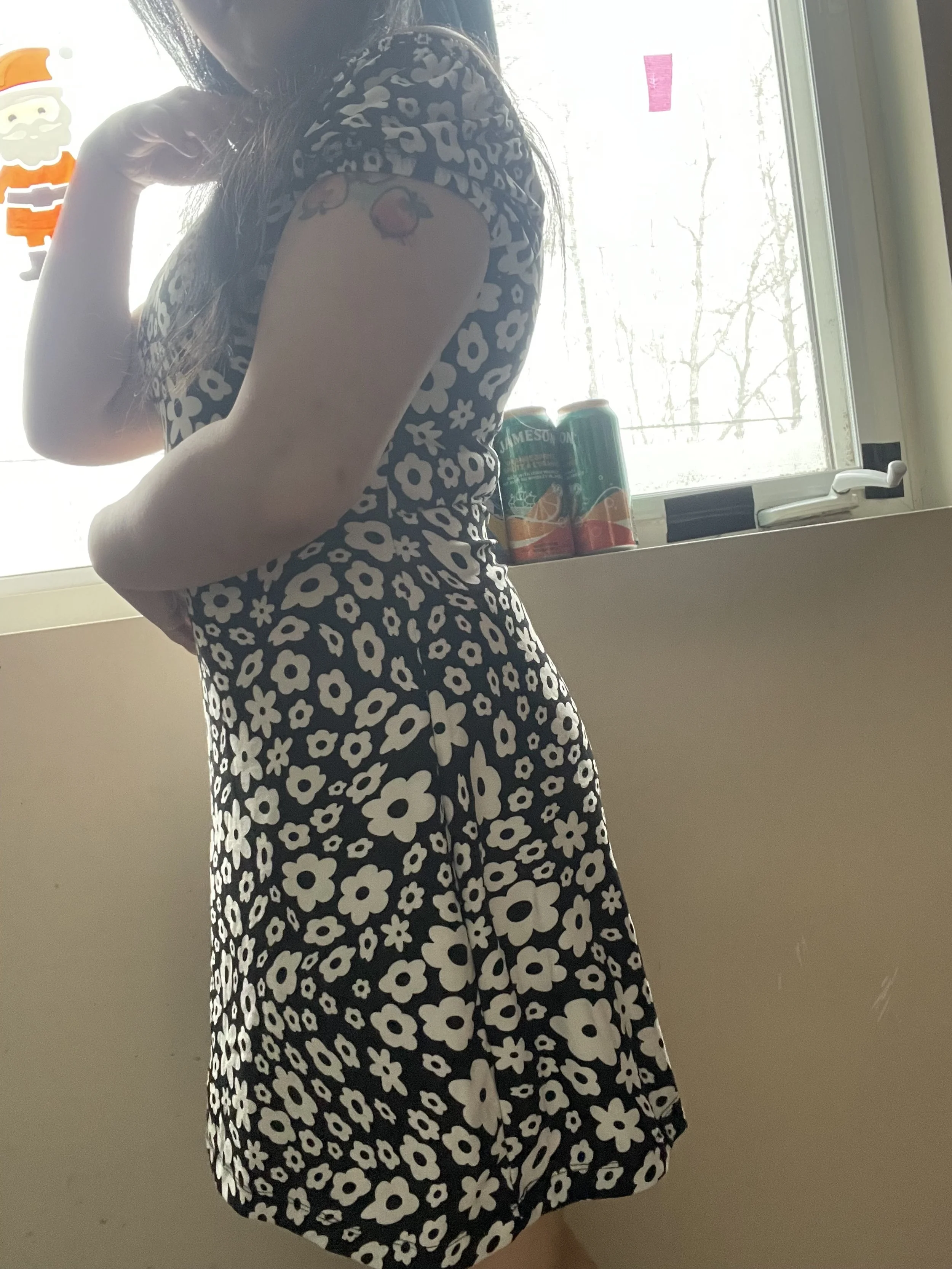 Woman wearing a black and white floral dress, showing a tattoo of a peach on her upper arm, standing near a window with a snow-covered scene outside.