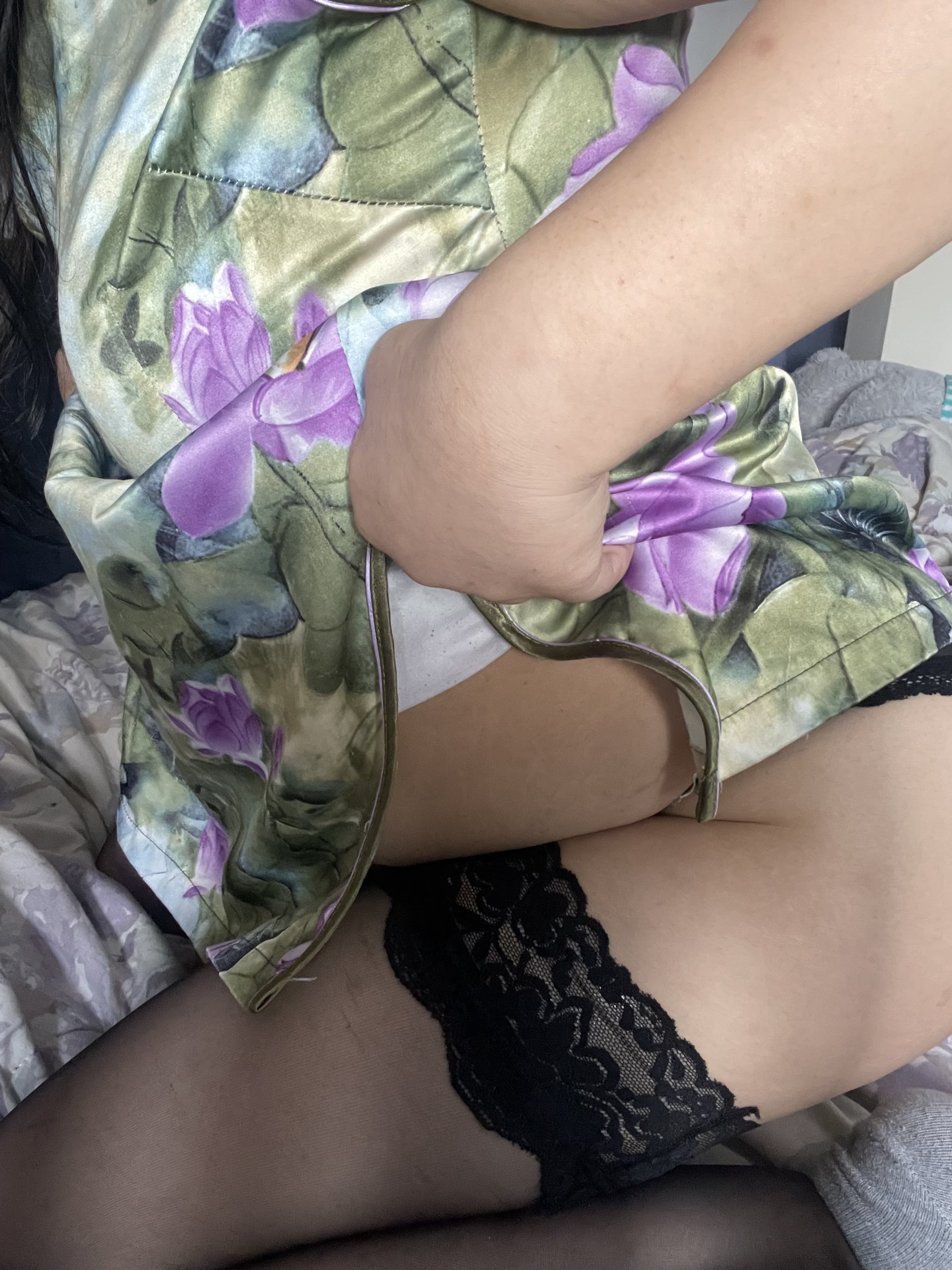 A person wearing black lace stockings and a floral satin robe, with one hand in the robe opening, sitting on a bed.