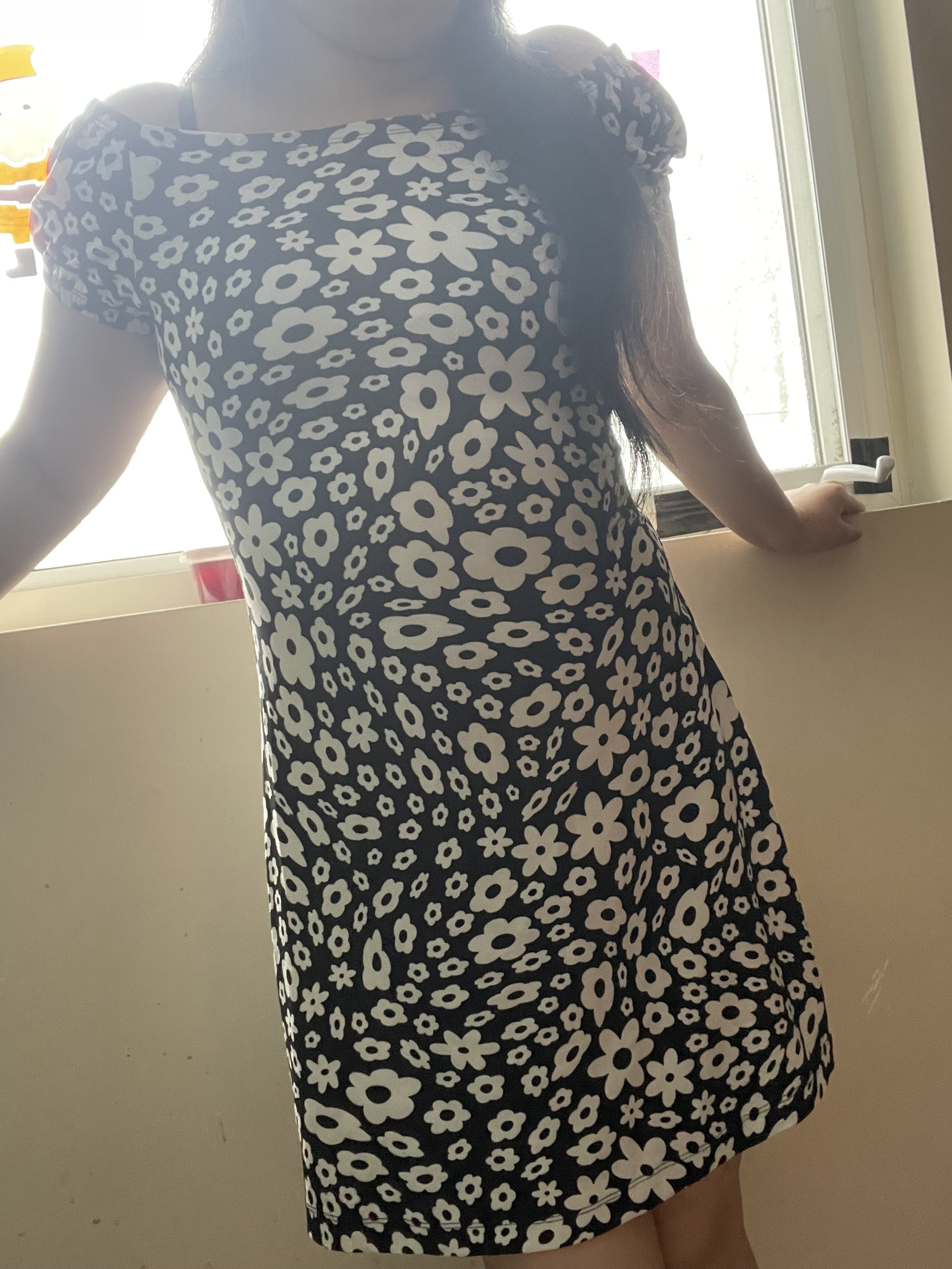Person wearing a black and white floral dress, leaning on a windowsill, with sunlight coming through the window.