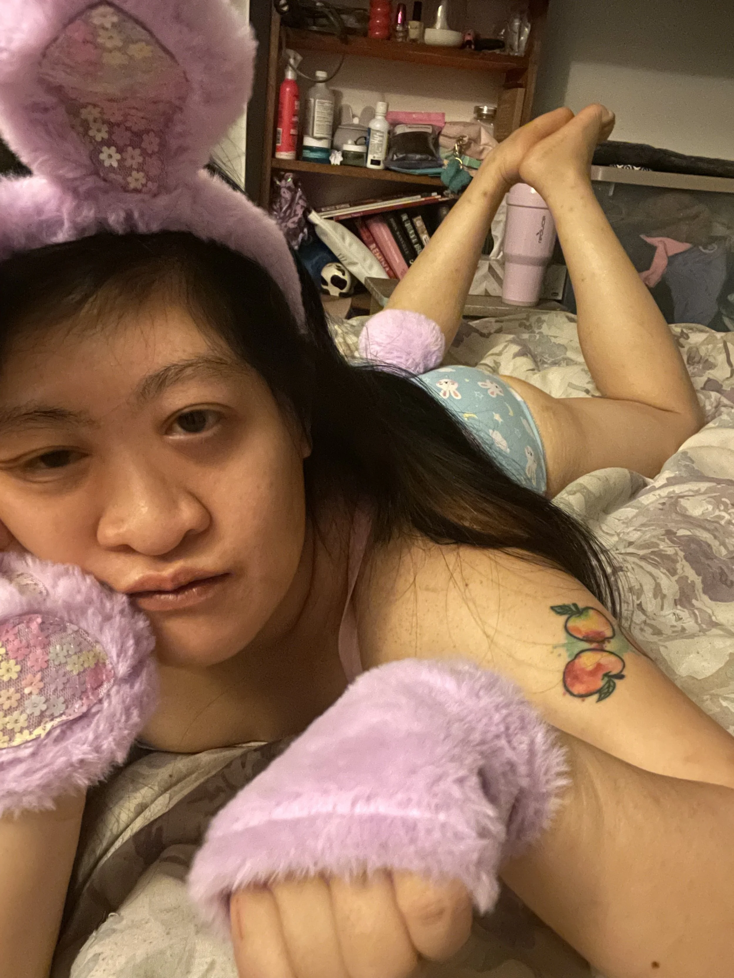 A woman lying on a bed with pink fuzzy bunny slippers and a pink bunny headband. She has a tattoo of two peaches on her upper arm and is wearing blue pajama shorts with floral pattern. The background shows a room with a bookshelf, various bottles, and a pink tumbler.