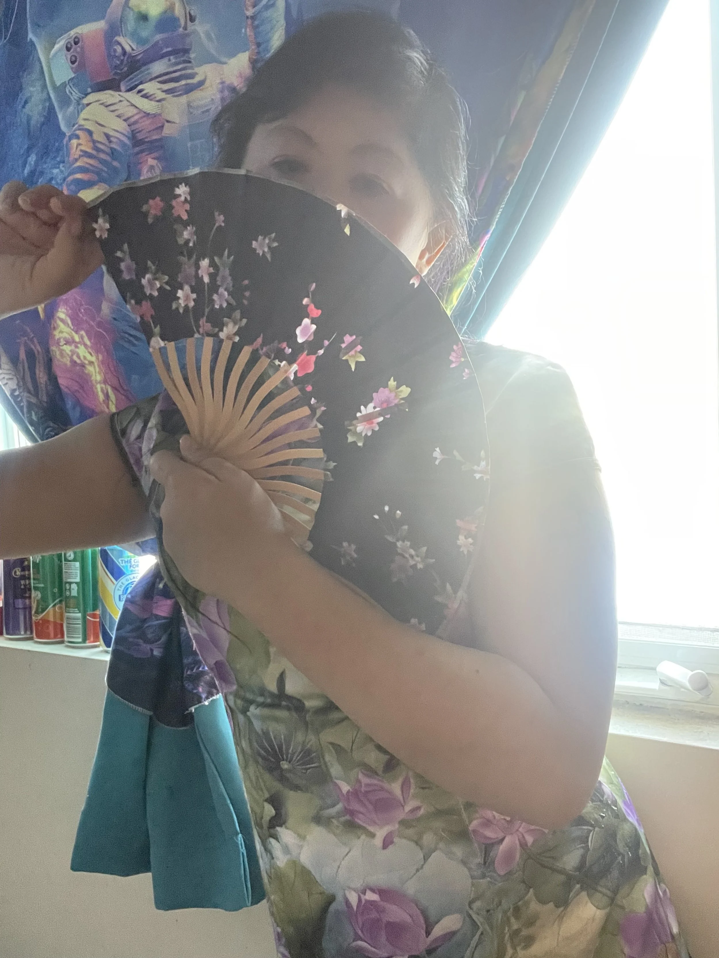 Woman holding a floral-patterned folding fan partially covering her face, indoors near a window with bright light, colorful wall art, and cans on a shelf in the background.