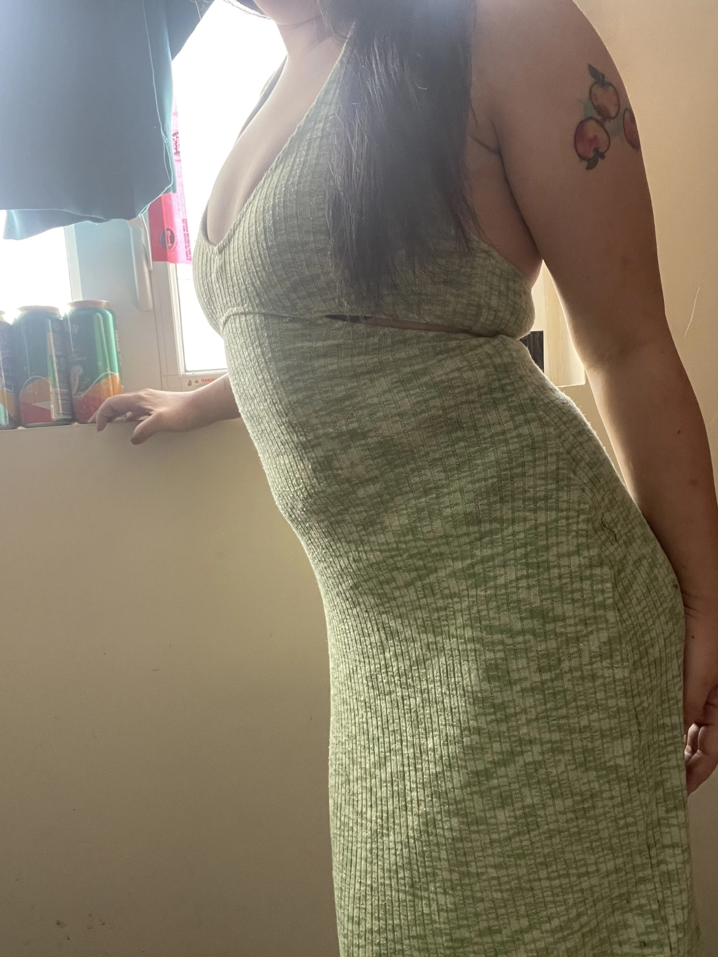 A woman wearing a green tank top with a cut-out detail, showing a tattoo of peaches on her upper arm, is standing near a window with sunlight coming through. The window has cans of soda and a blue cloth hanging nearby.