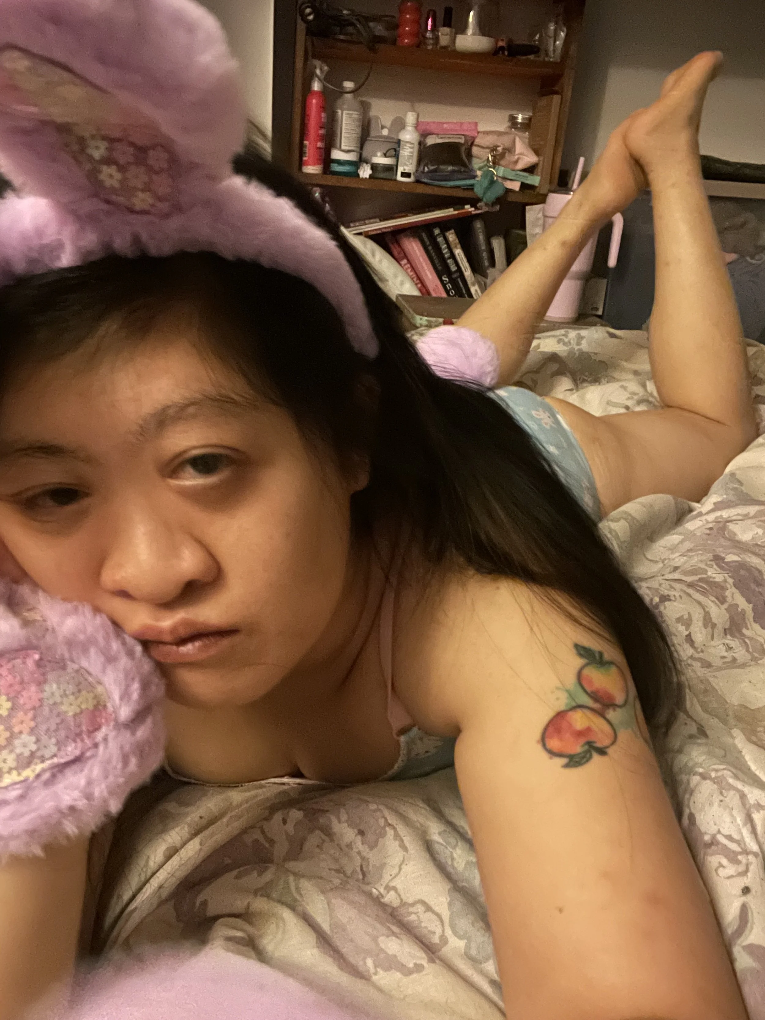 A young woman with a peach tattoo on her upper arm is lying on a bed, resting her head on a pink plush pillow or stuffed animal. She has long dark hair and is wearing a light-colored tank top. The background shows a bookshelf with various bottles and